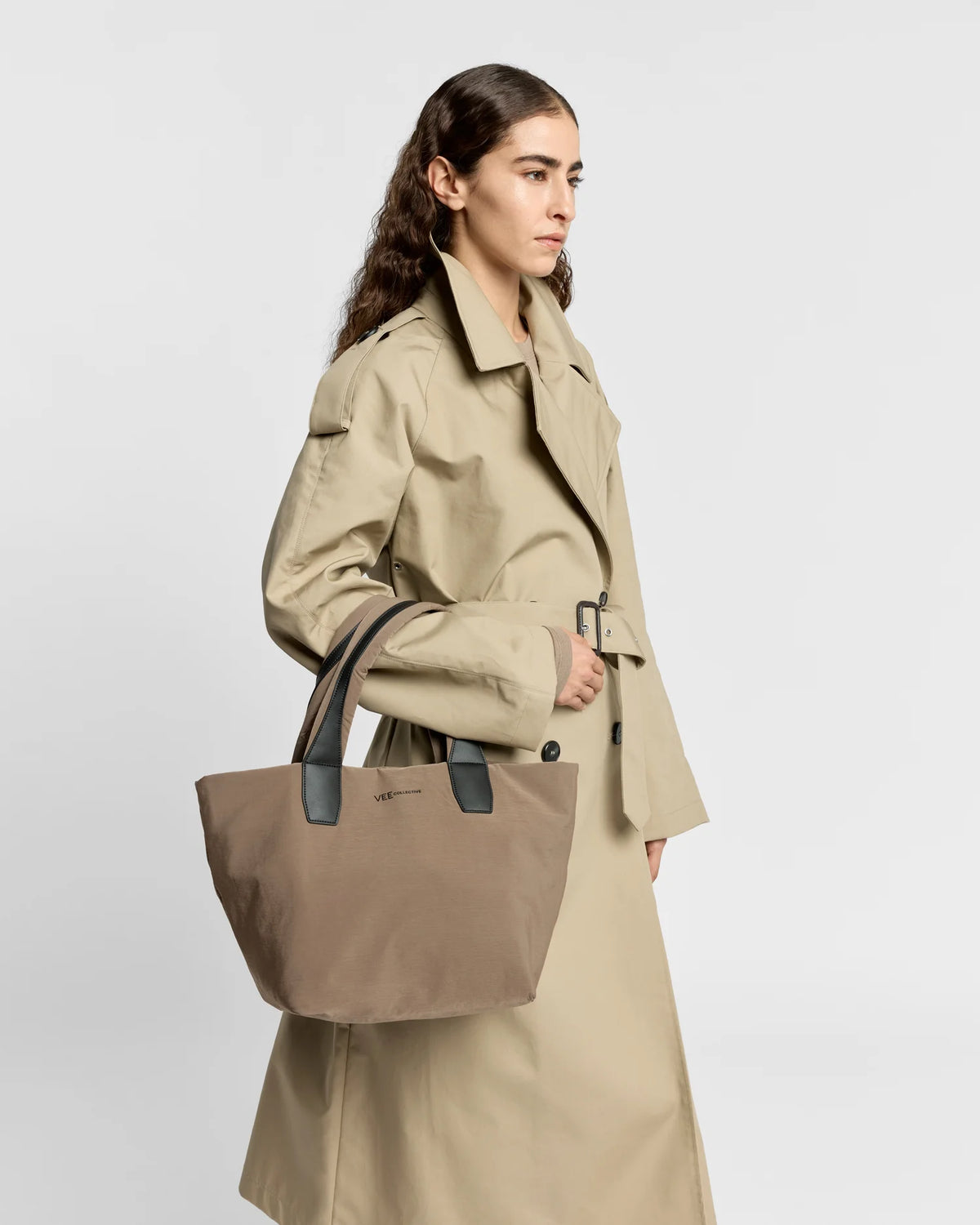 Woman in a beige trench coat carrying the Porter Essence Small Tote Bag in Mink Trench by Vee Collective against a plain studio background.