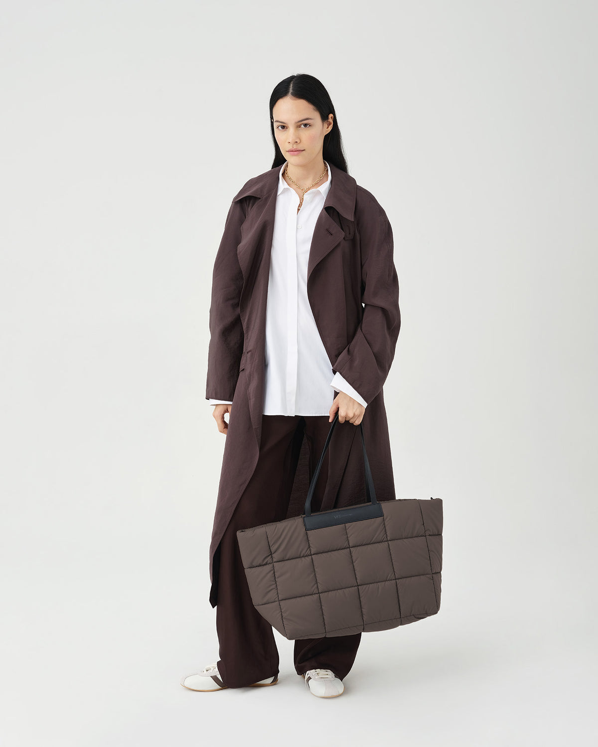 A photo of a model wearing a dark brown double-breasted trench coat over a white shirt and dark brown trousers, holding a mink Porter Marche bag against a white studio background.
