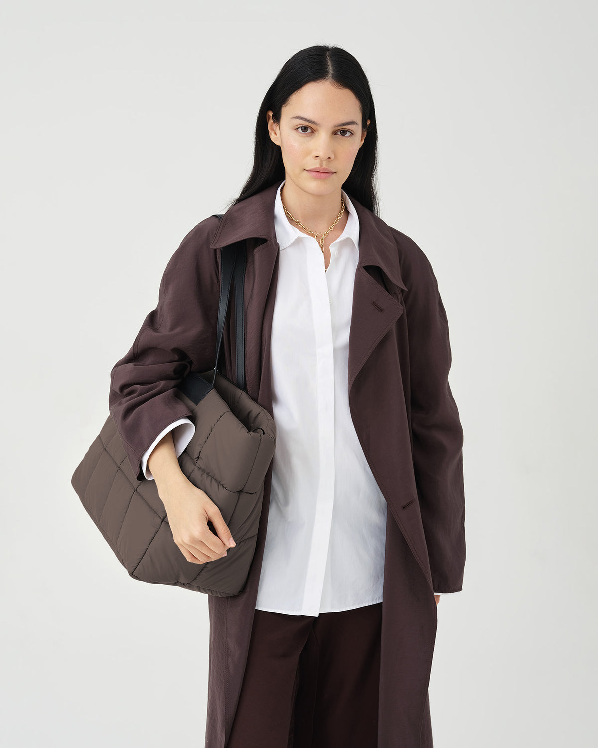A photo of a model wearing a dark brown double-breasted trench coat over a white shirt and dark brown trousers, holding a mink Porter Weekender bag against a white studio background.