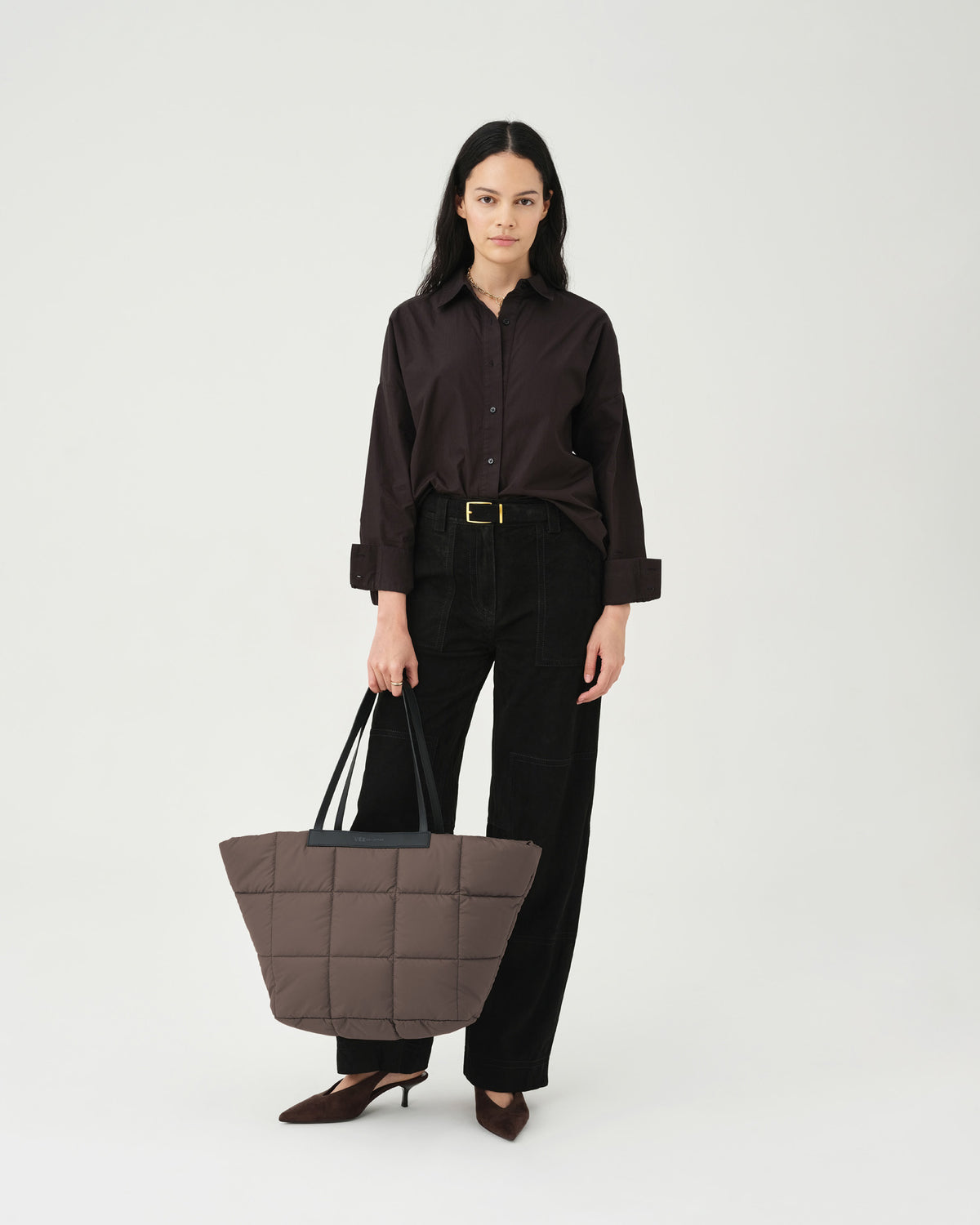 A model stands against a light backdrop, holding a small quilted tote bag in a soft brown color. She wears a dark button-up shirt and black trousers, showcasing the Porter marche mink with square patterns.