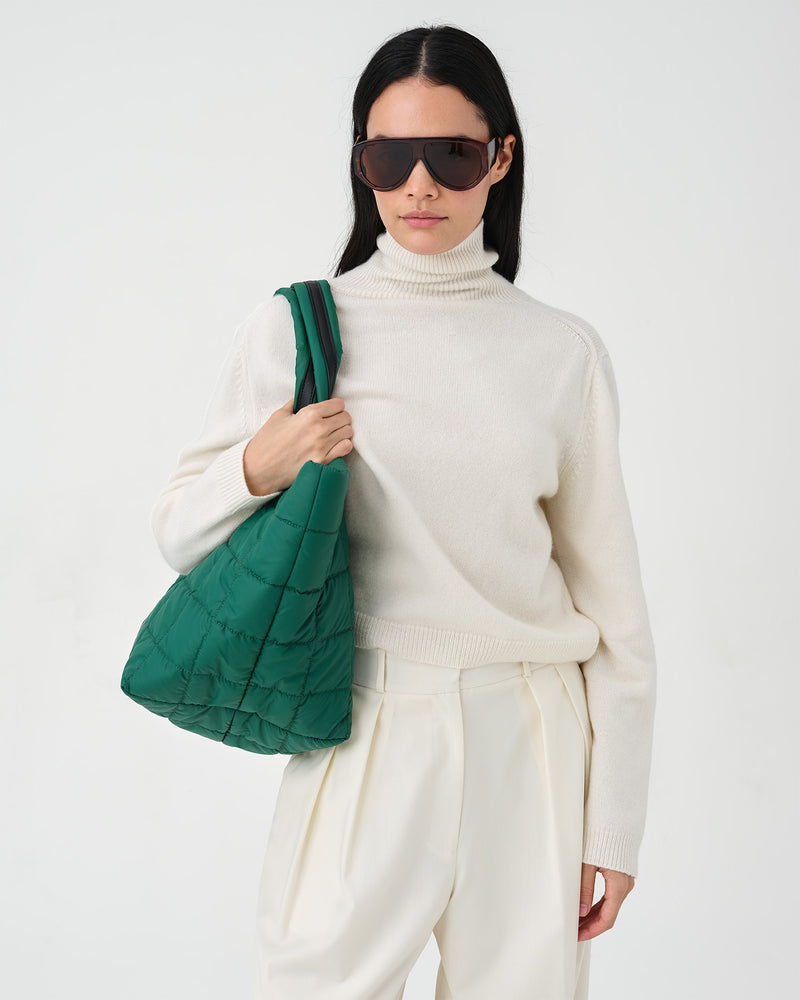 A model wearing a cream turtleneck sweater and matching cream trousers, styled with dark sunglasses and a green quilted shoulder bag, against a plain white studio background.