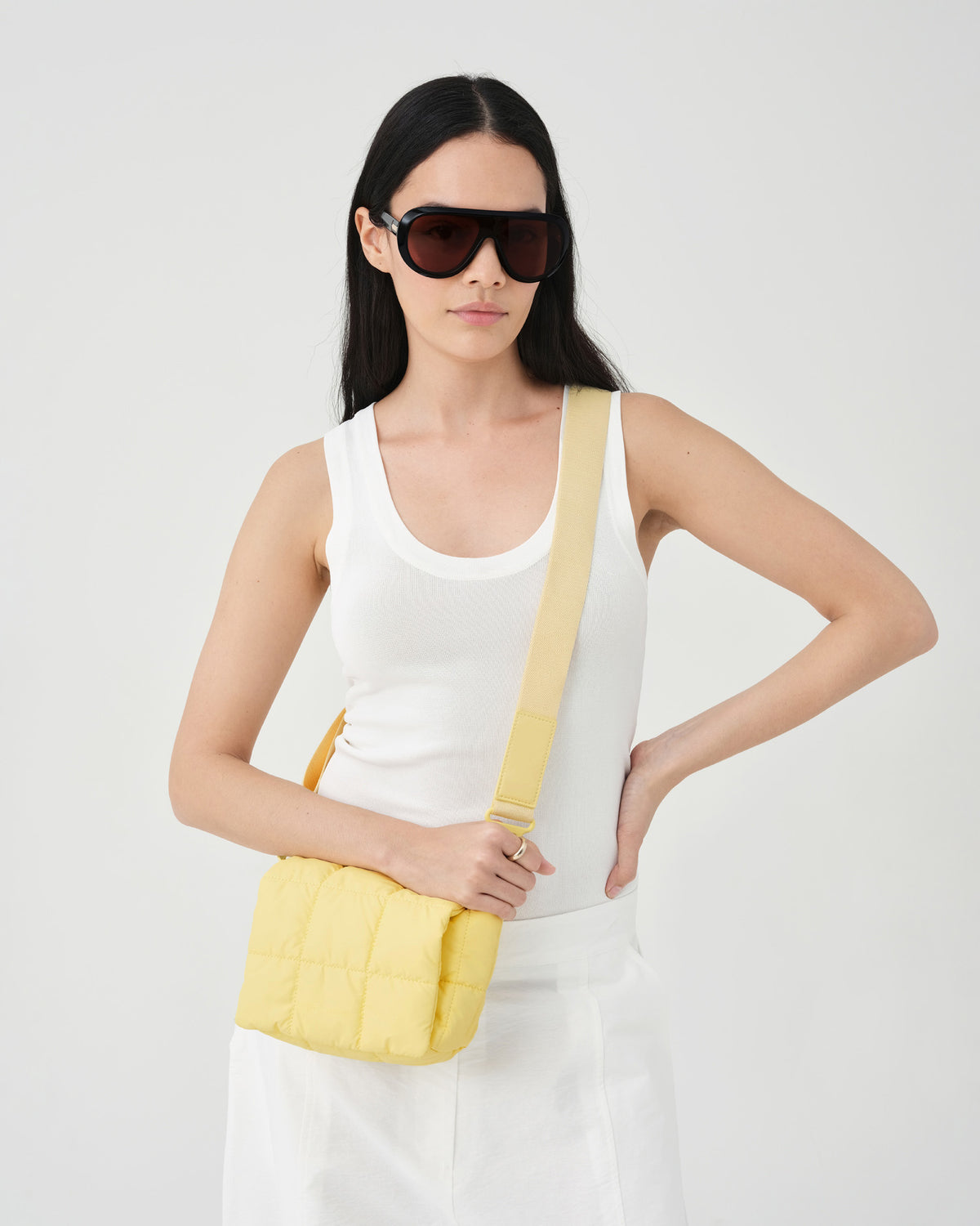 A photo of a woman wearing a white sleeveless top and white wide-leg pants, carrying a buttermilk Porter Messenger shoulder bag against a plain light gray studio background.