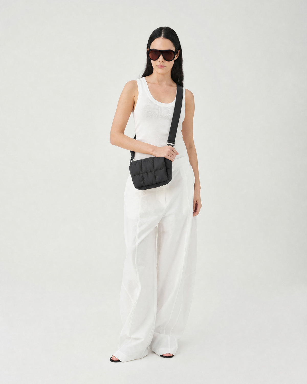 A model wearing a white tank top and white wide-leg trousers, accessorized with a black quilted crossbody bag and black sunglasses, standing against a plain white studio background.