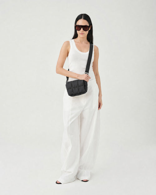 A model wearing a white tank top and white wide-leg trousers, accessorized with a black quilted crossbody bag and black sunglasses, standing against a plain white studio background. c__2