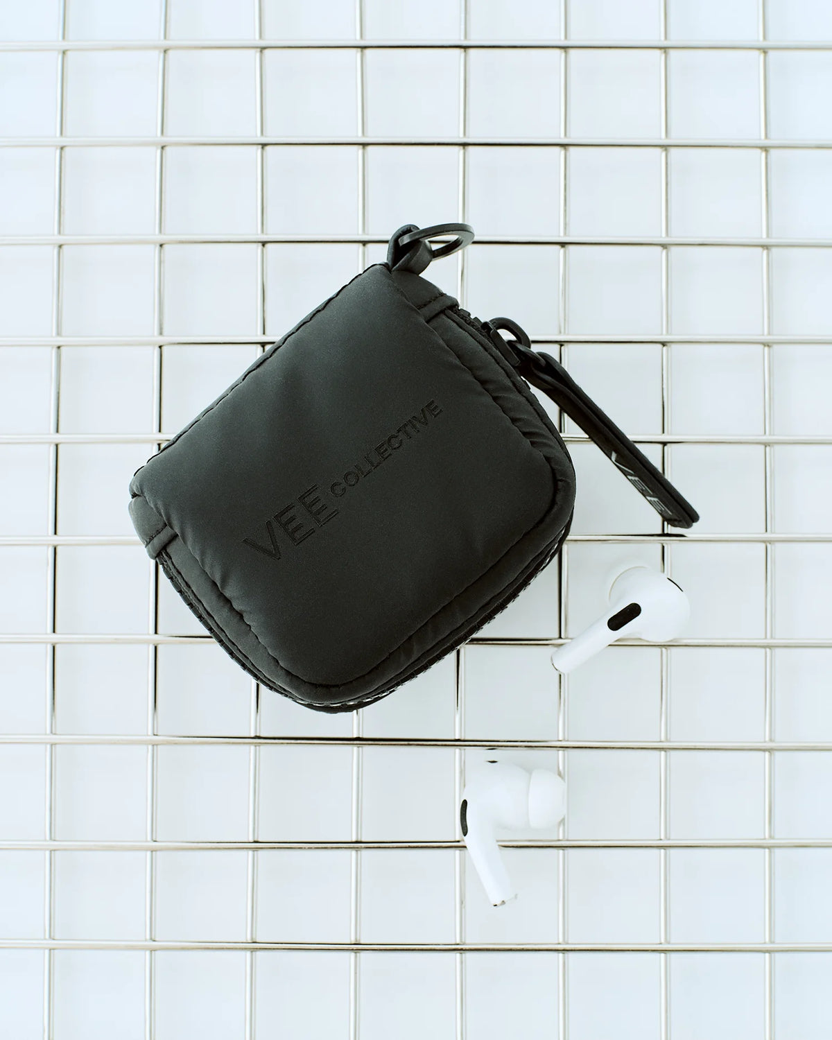 A Matt Black Vee Collective Porter Pods Case resting on a metal wire grid, shown with two white wireless earbuds placed nearby.