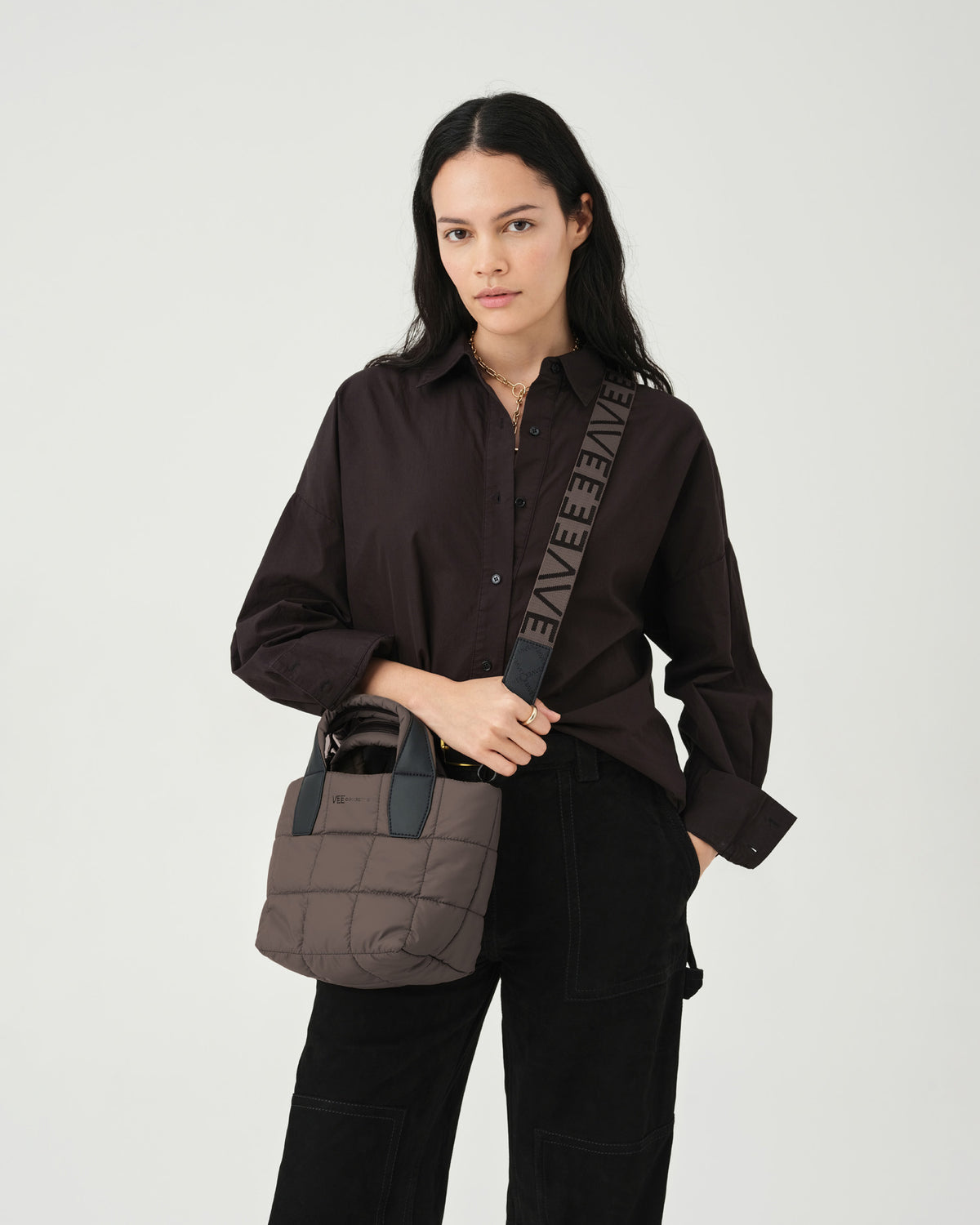 A model stands against a light backdrop, holding a small quilted tote bag in a soft brown color. She wears a dark button-up shirt and black trousers, showcasing the Porter totes mink square pattern and comfortable shoulder straps.