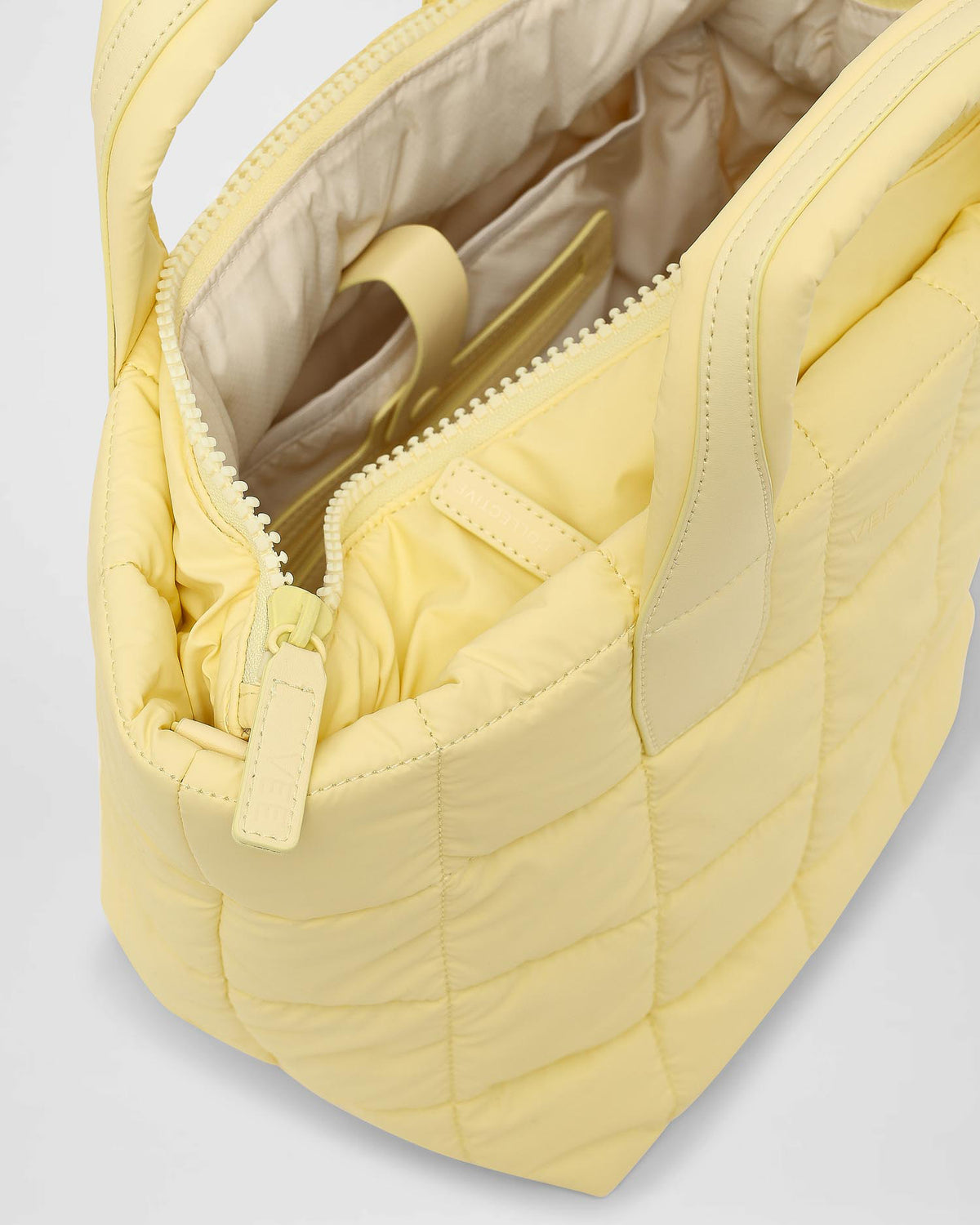 An overhead view of a small quilted tote bag in a soft buttermilk yellow color, featuring puffy square patterns. The bag is partially open, revealing a beige interior with pockets and a zipper closure.