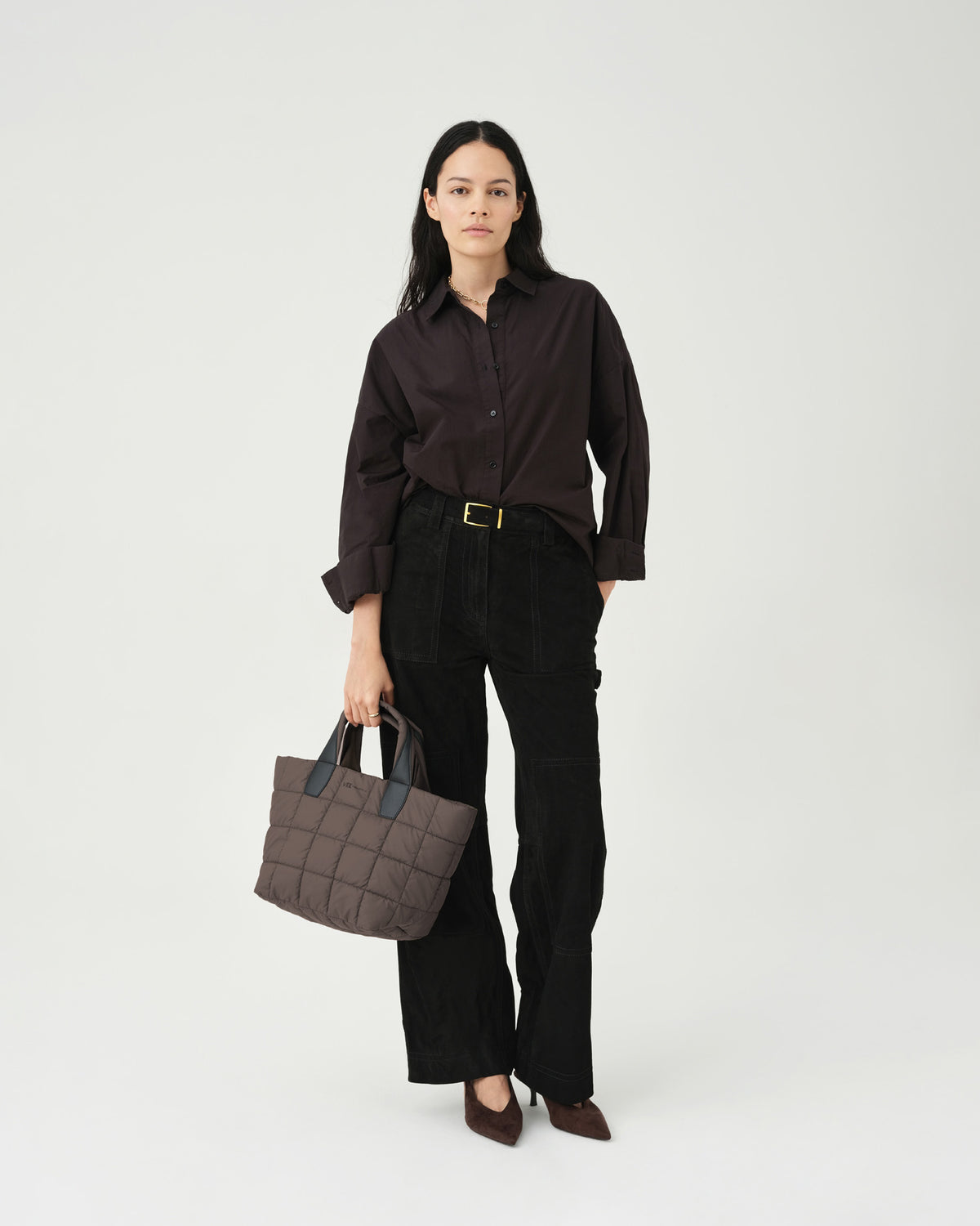 A model stands against a plain white background, wearing a dark oversized shirt and black high-waisted pants. She holds a small quilted tote bag in a mink color, complemented by pointed-toe heels, showcasing a stylish, casual look.