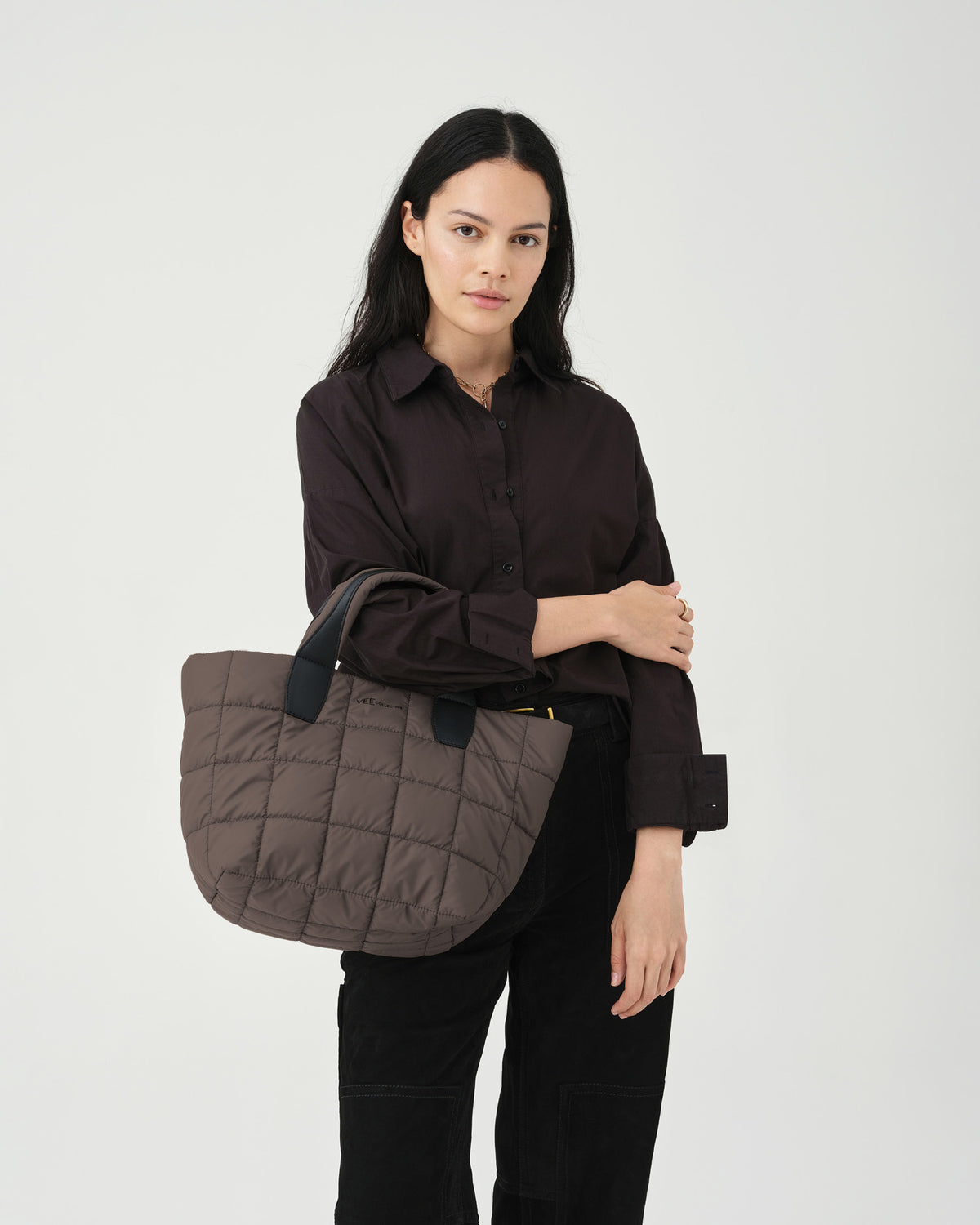 A model stands against a light backdrop, holding a small quilted tote bag in a soft brown color. She wears a dark button-up shirt and black trousers, showcasing the Porter totes mink square pattern.