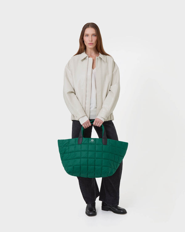 A model stands against a light gray studio background holding a large quilted green tote bag with black handles. The model is wearing a light beige jacket over a white top, dark wide-leg pants, and black shoes. c__2
