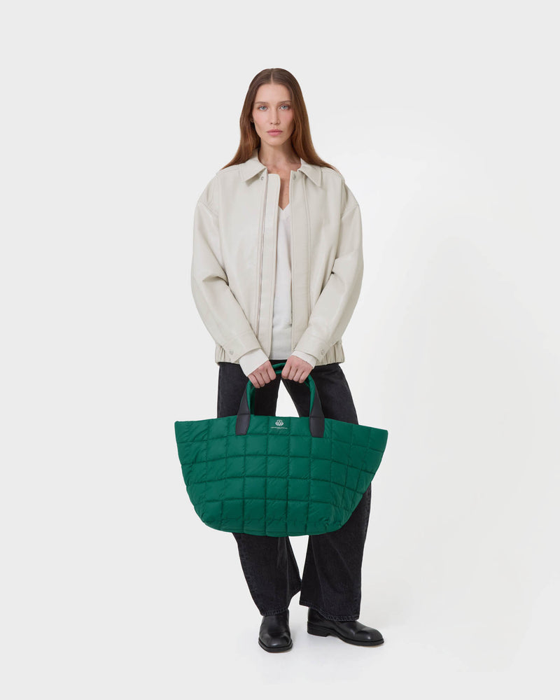 A model stands against a light gray studio background holding a large quilted green tote bag with black handles. The model is wearing a light beige jacket over a white top, dark wide-leg pants, and black shoes.