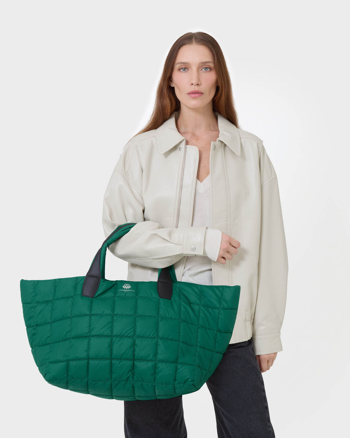 A woman modeling a large quilted green tote bag with black handles, styled with a cream jacket and dark pants against a light gray studio background.