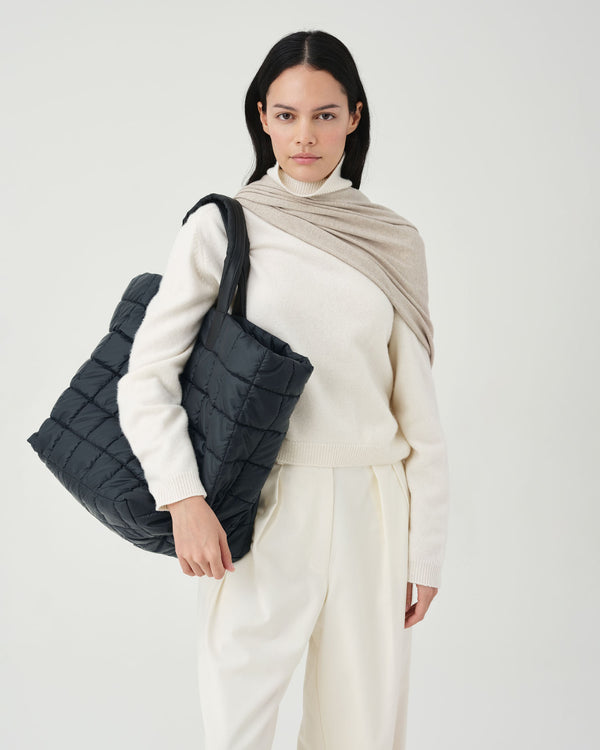 A model wearing a cream knit turtleneck and matching trousers, styled with a beige shawl, carrying a porter weekender bag against a  white studio background
