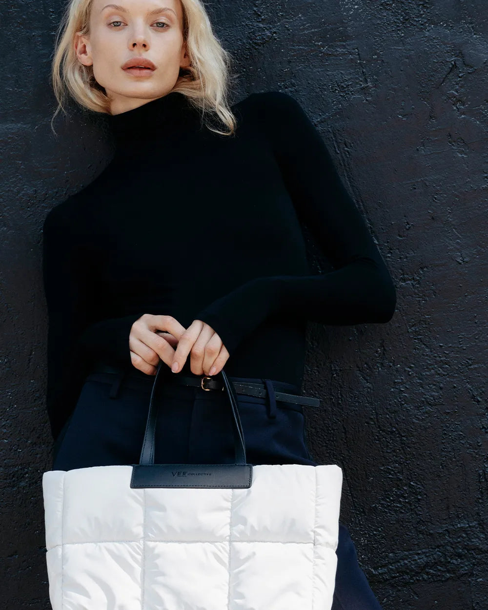 Model wearing a black turtleneck and holding a white quilted tote bag against a textured black wall.
