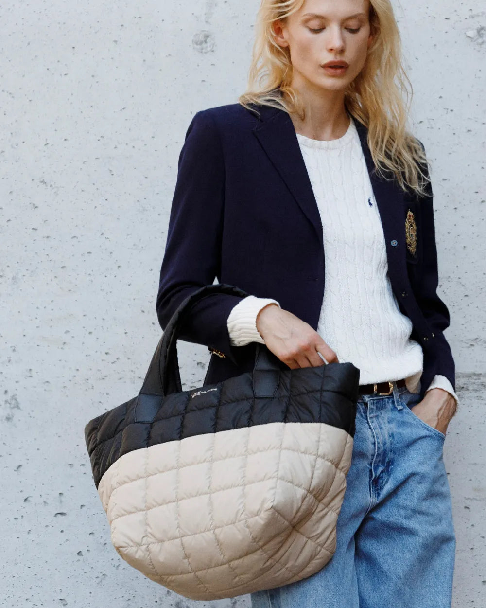 Fashionable woman in a blazer holds a Porter Tote Medium in Black Desert against a textured wall.