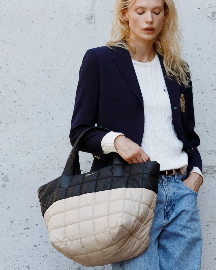 Fashionable woman in a blazer holds a Porter Tote Medium in Black Desert against a textured wall.