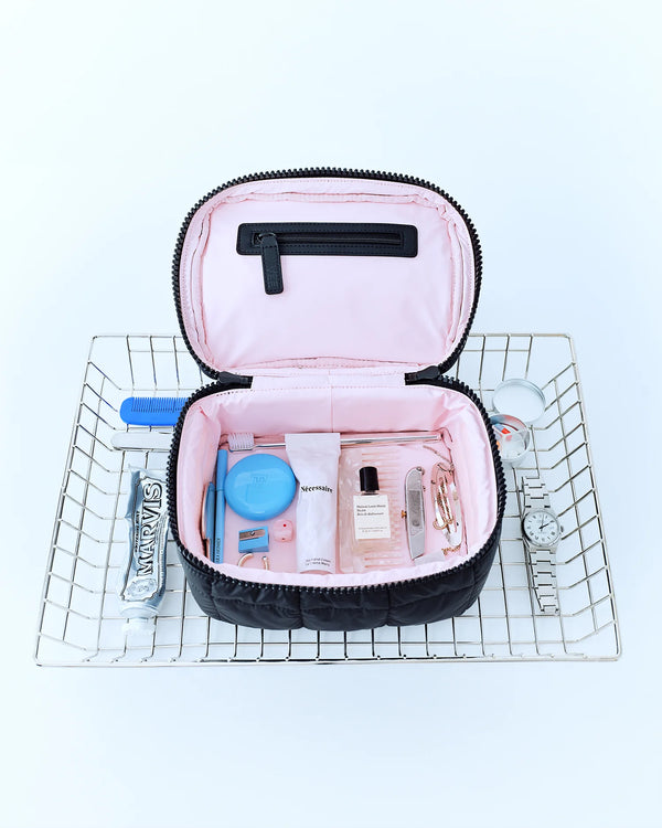 An open Porter Vanity Case by Vee Collective in Matt Black with a light pink interior, filled with grooming items such as a toothbrush, comb, skincare products, perfume, and more.