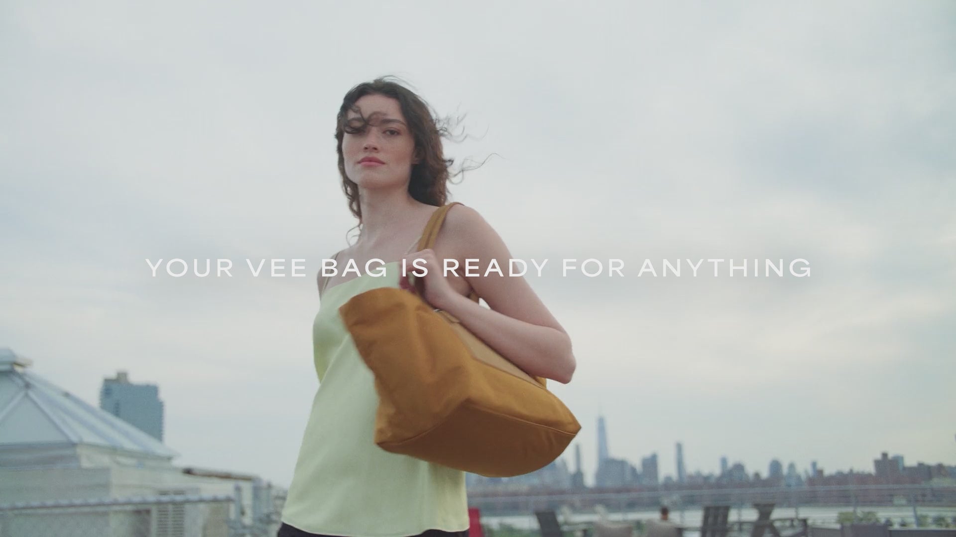 A woman with brown hair walking with the Duo Tote Large Amber bag. In the middle, the sentence “Your Vee bag is ready for anything” is written.

