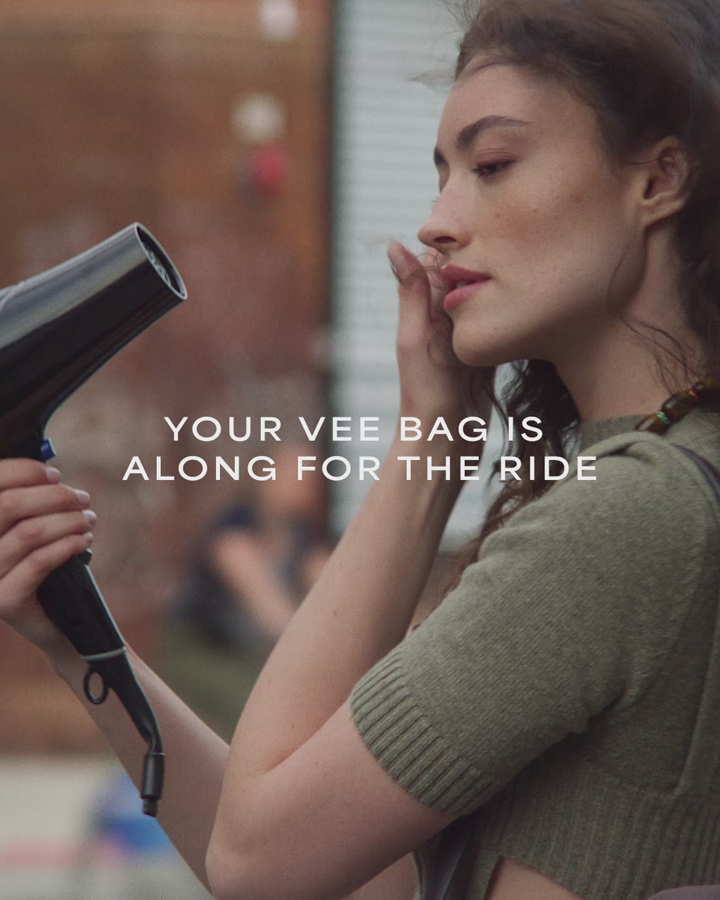 A video featuring a model drying her hair with a blow dryer while carrying a Vertical Tote Shale Bag, with a brick house visible in the background.