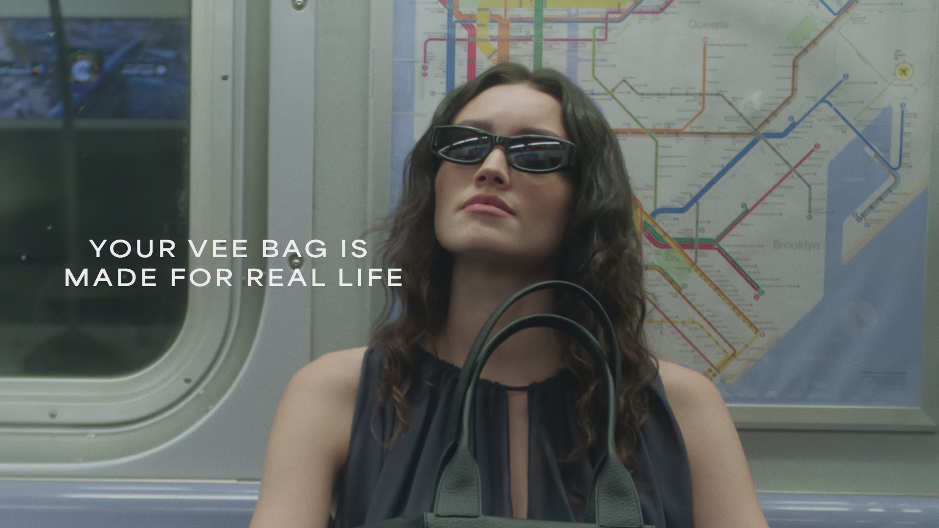 A video featuring a model wearing black sunglasses and a sheer black sleeveless top aboard a train, carrying a large black Duo tote bag with a leather front pocket.