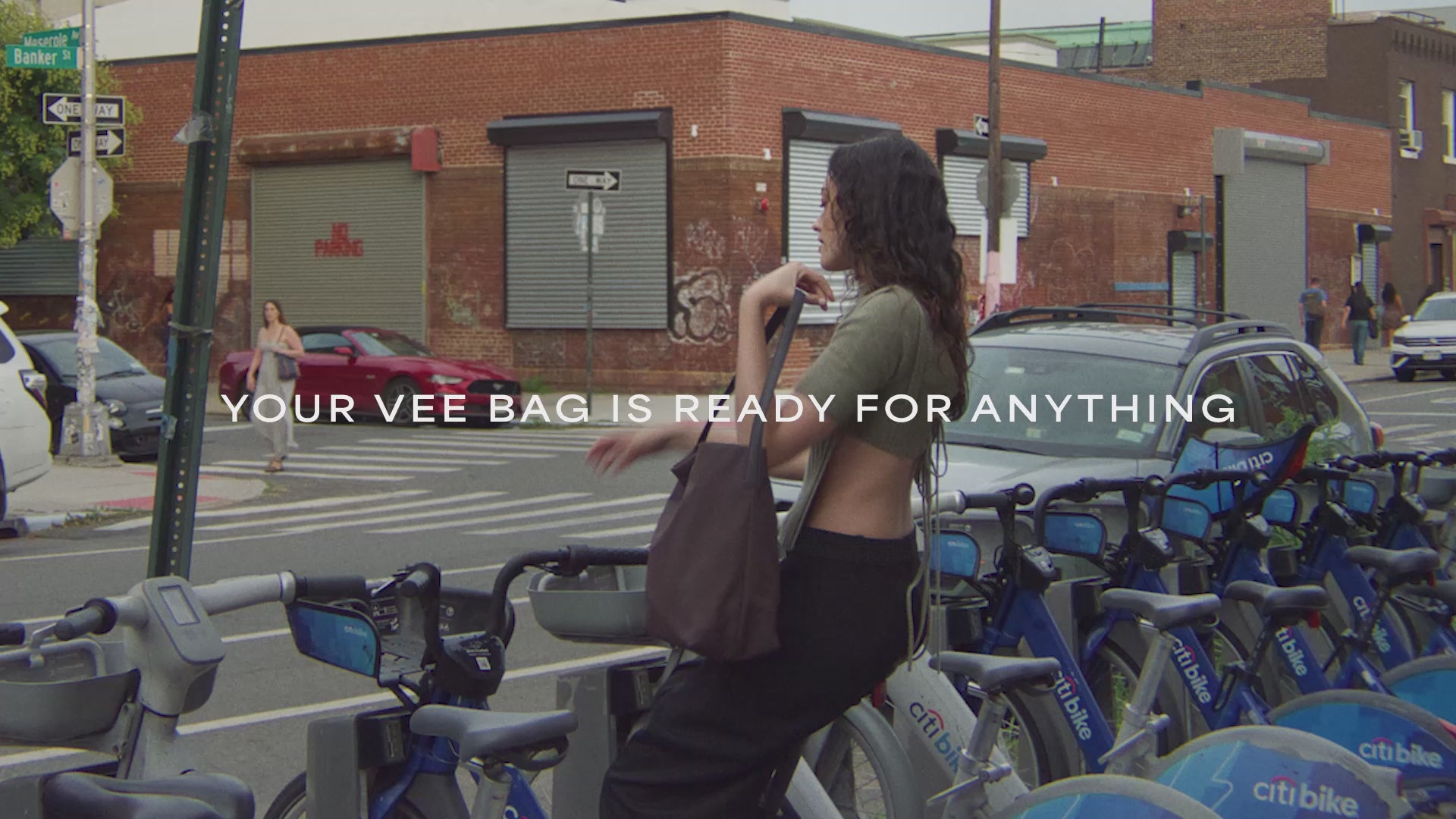 A video featuring a model getting off a bicycle while carrying a Vertical Tote Shale Bag, with a cars and brick house visible in the background.
