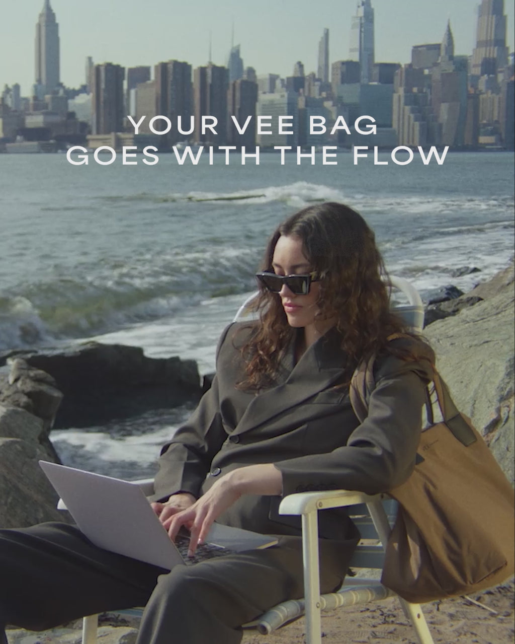 A video featuring a model wearing black sunglasses and a jacket, carrying an Essence Medium Mink Bag while seated on a chair by the riverside in the city.