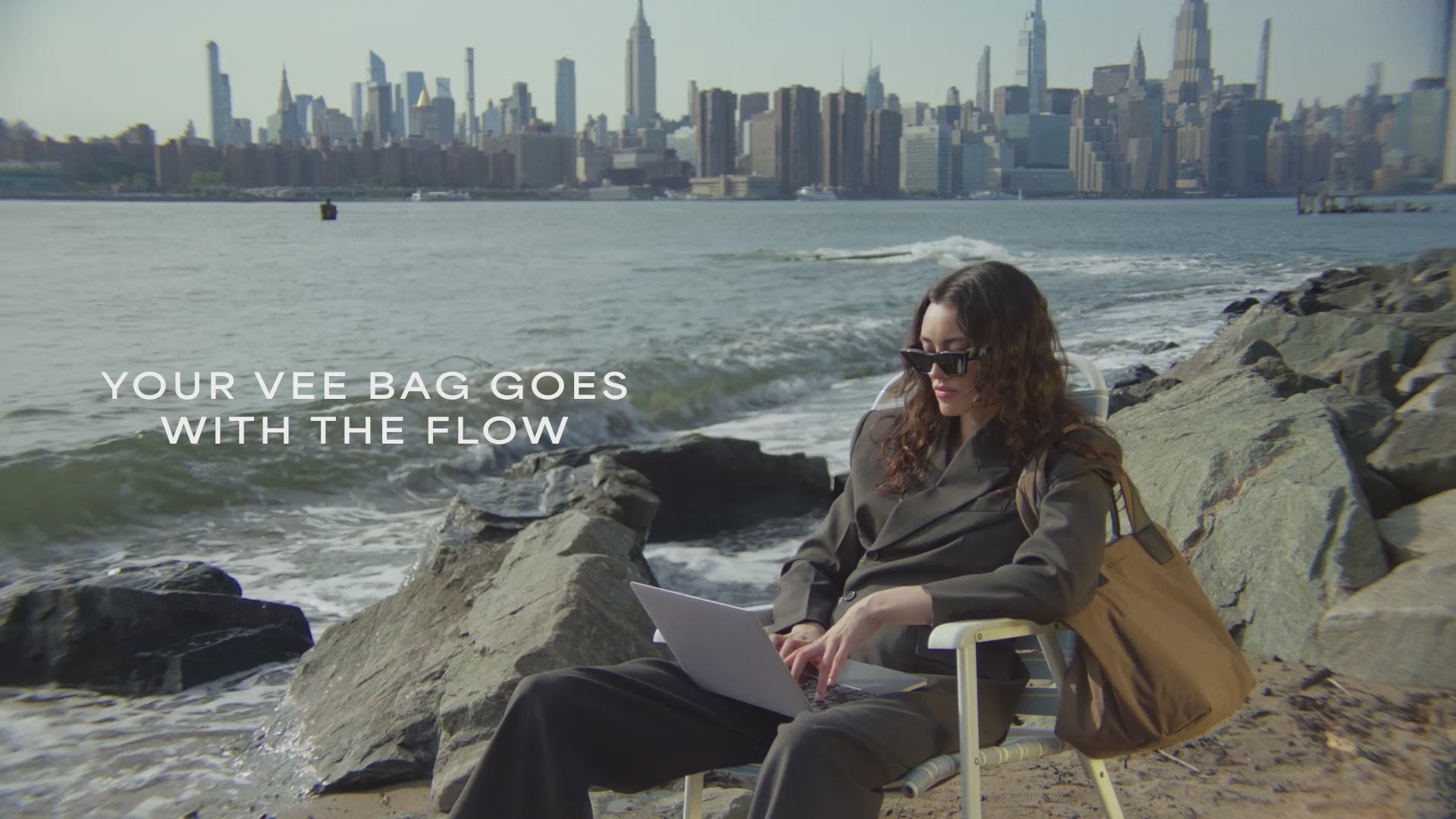 A video featuring a model wearing black sunglasses and a jacket, carrying an Essence Medium Mink Bag while seated on a chair by the beach in the city.