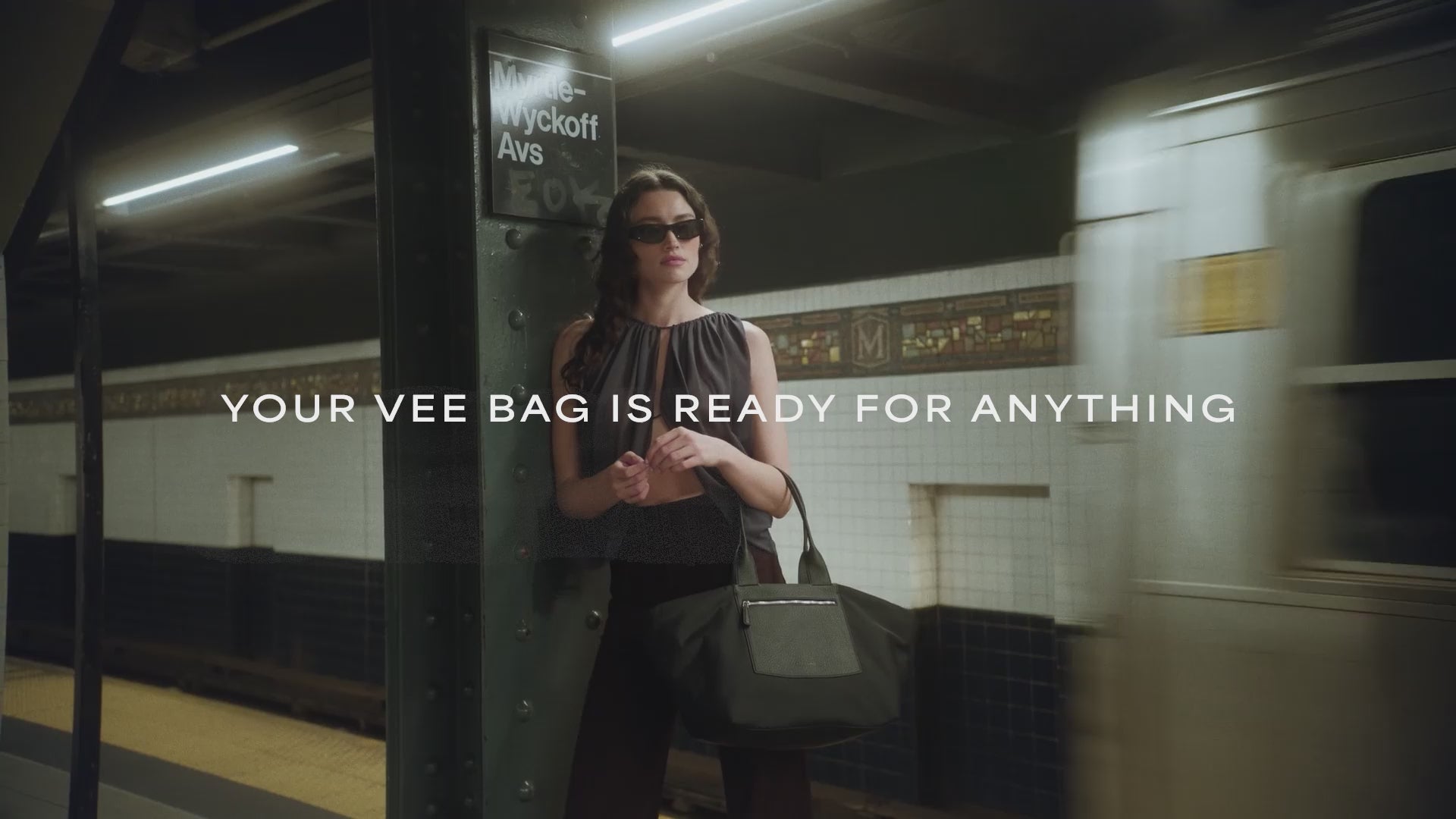 A video featuring a model wearing black sunglasses and a sheer black sleeveless top is seen at a train station and aboard a train, carrying a large black Duo tote bag with a leather front pocket and handles.