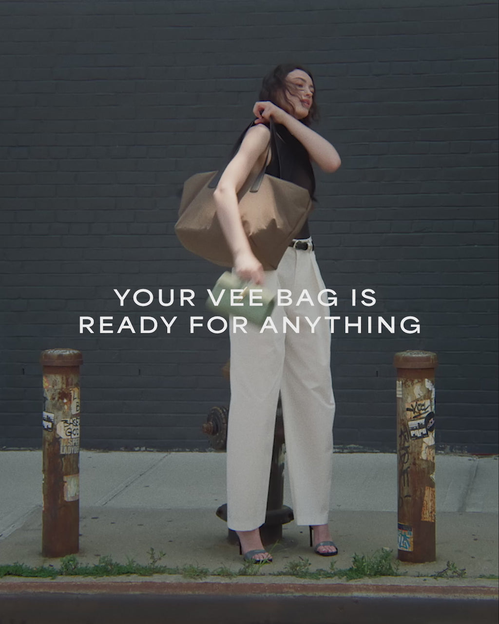 A video featuring a model wearing a sleeveless top and white trousers, carrying an Essence Medium Mink Bag while walking with a brick background.