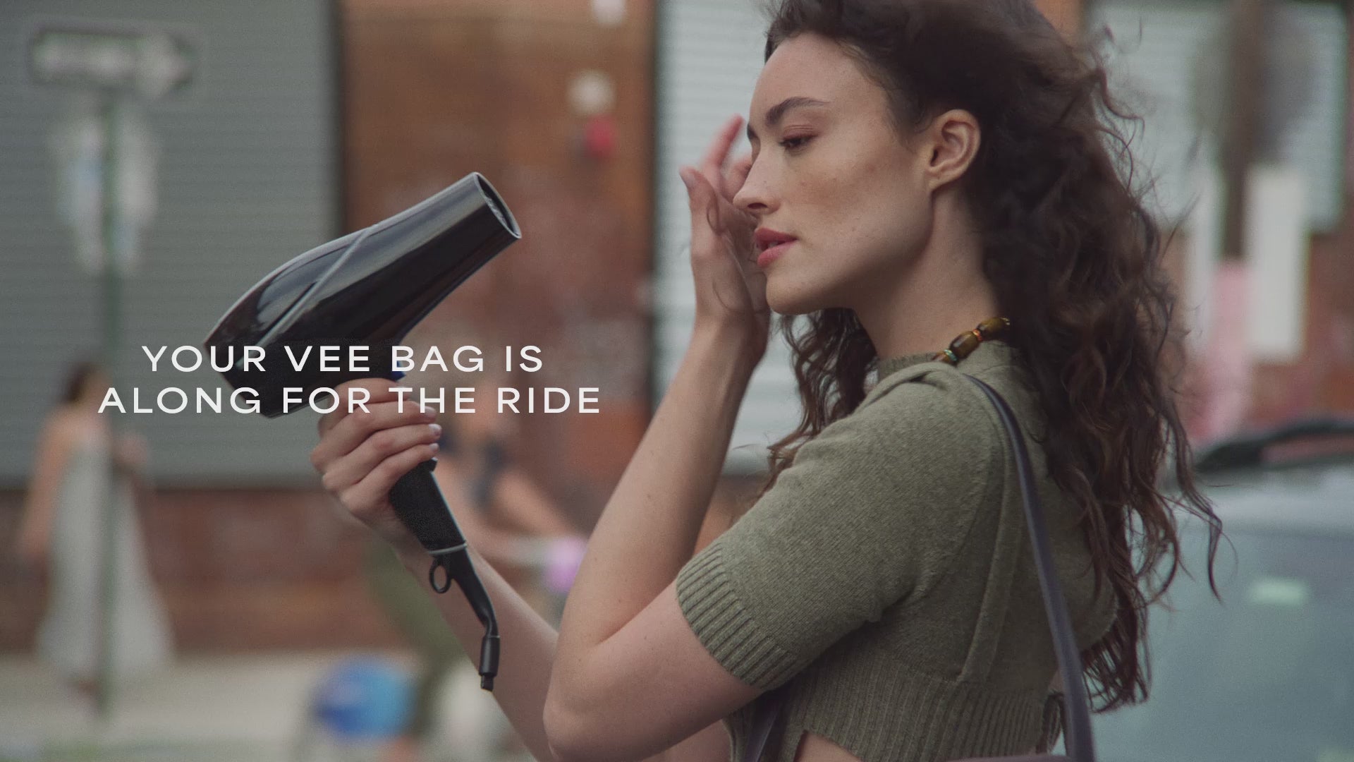 A video featuring a model drying her hair with a blow dryer while carrying a Vertical Tote Shale Bag, with cars and brick house visible in the background.