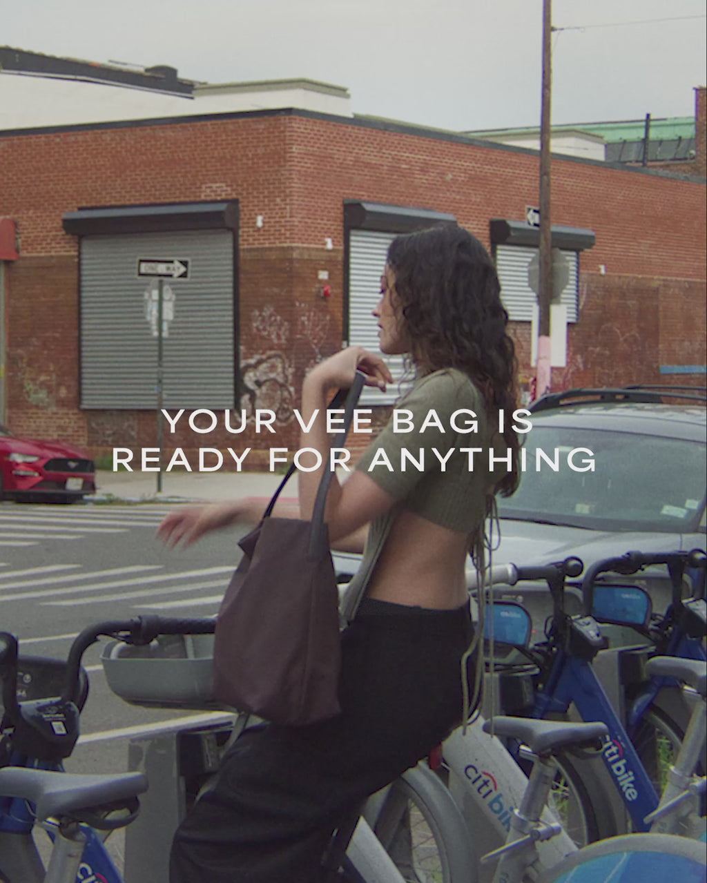 A video featuring a model wearing a sleeveless top and white trousers, carrying a Vertical Tote Shale Bag while walking, with a car and brick house visible in the background.
