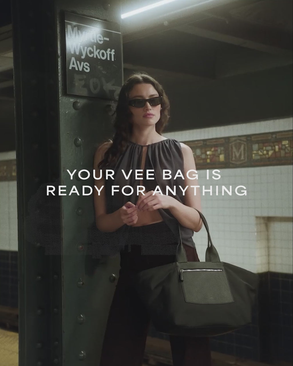 A video featuring a model wearing black sunglasses and a sheer black sleeveless top is seen at a train station and aboard a train, carrying a large black Duo tote bag with a leather front pocket and handles.