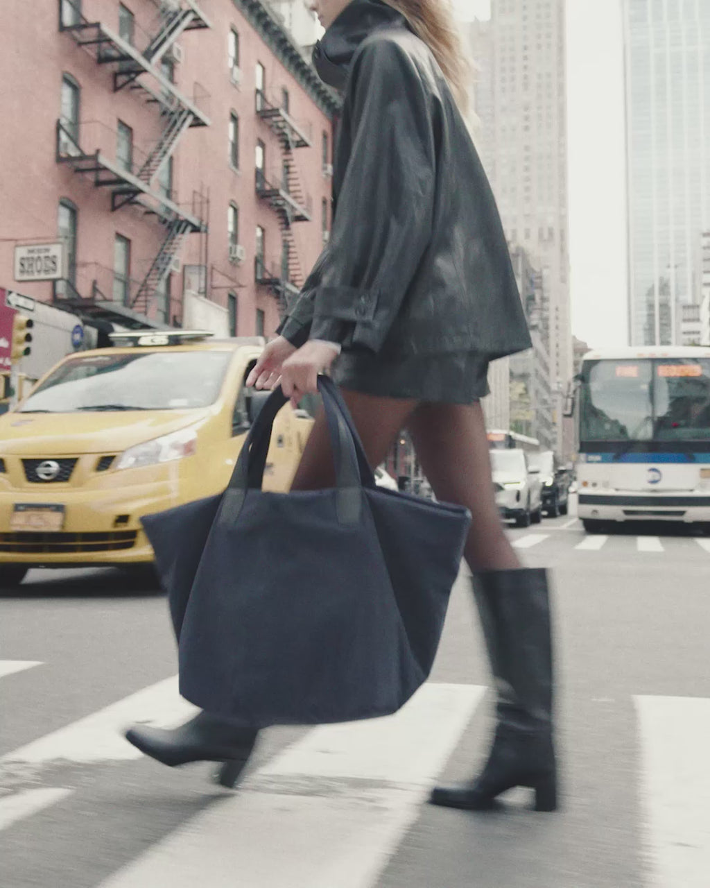 A woman walks on a city street wearing a black leather jacket and skirt, carrying Vee Collectives Porter Essence Weekender Tote Bag in Midnight Trench.