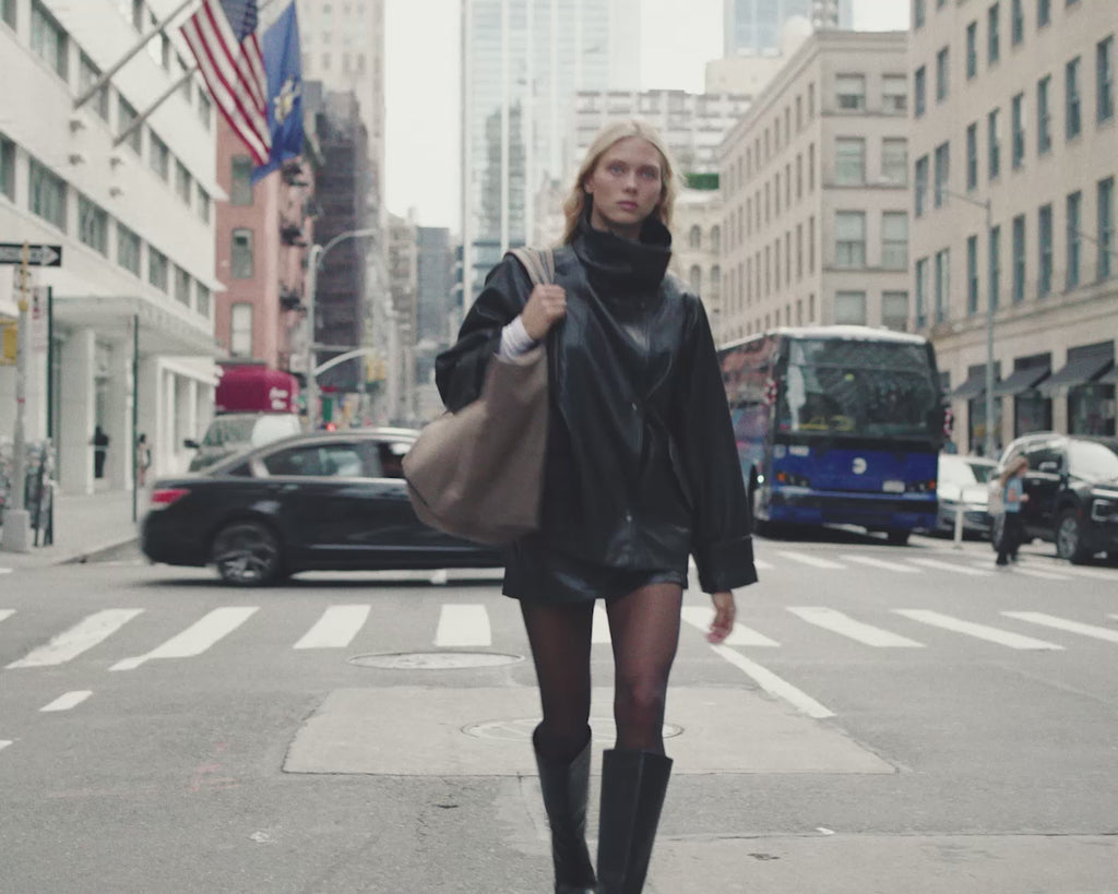 A woman with long blonde hair walks on a city street wearing a black leather jacket and skirt, carrying Vee Collectives Porter Essence Weekender Tote Bag in Mink Trench over her shoulder.
