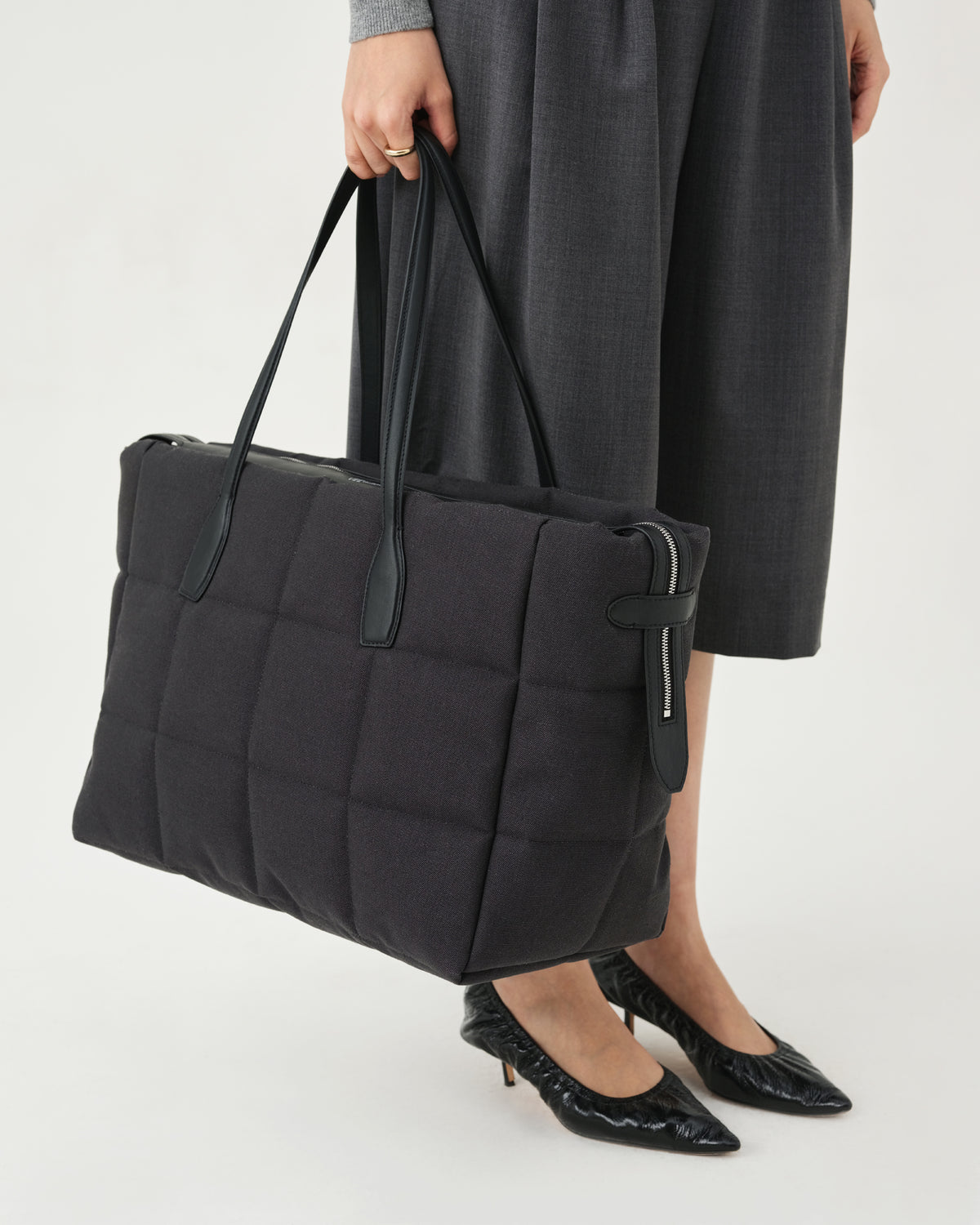 A photo of a model standing against a white studio background, wearing a gray long-sleeve knit top, dark charcoal pleated midi pants, and black pointed-toe heels, while carrying a Tela Tote with black handles.