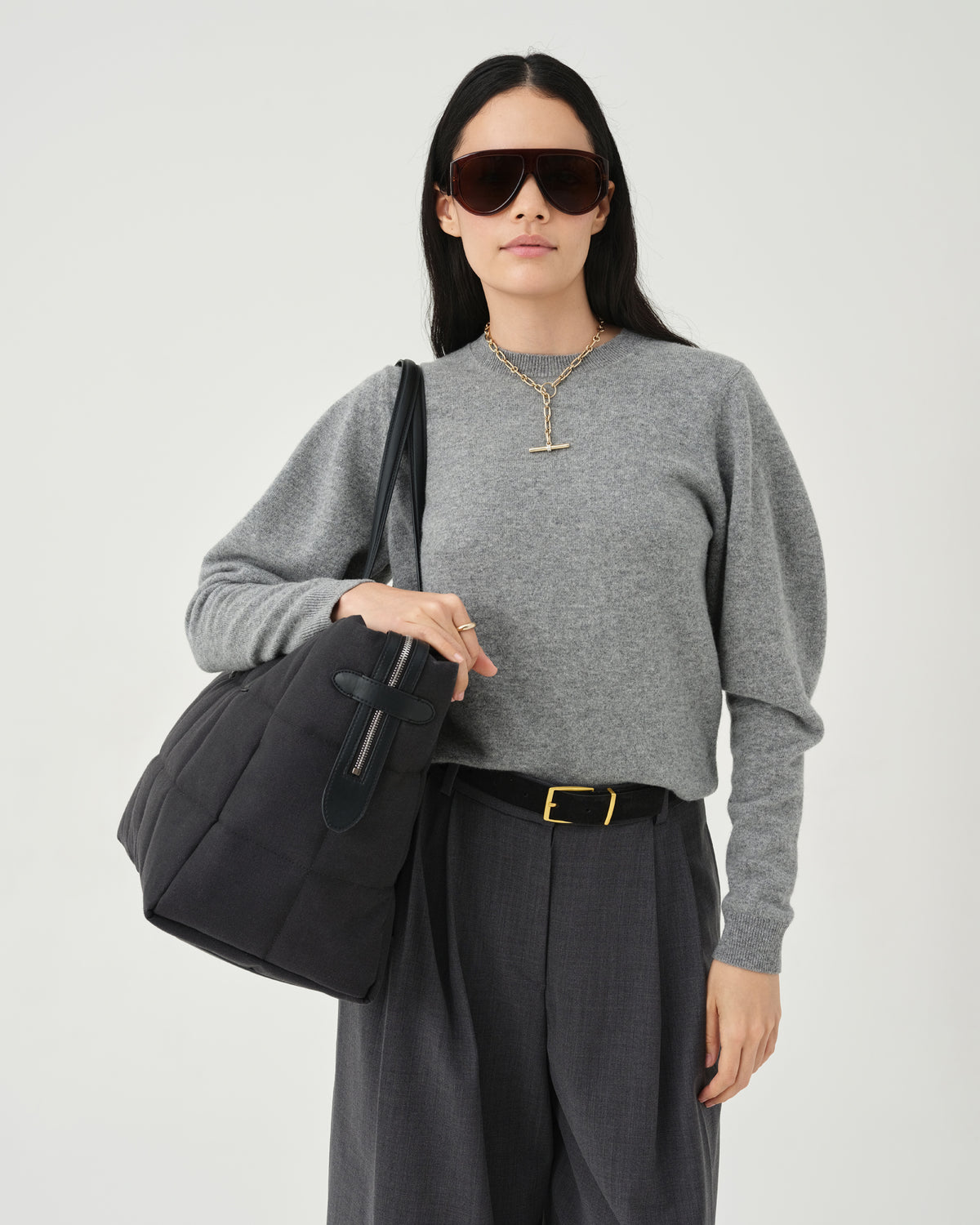 A photo of a model standing against a white studio background, wearing a gray long-sleeve knit top, dark charcoal pleated midi pants, while carrying a Tela Tote with black handles.