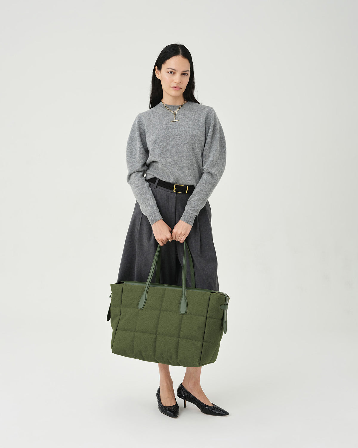 A photo of a woman modeling an olive Tela Tote bag with a matching shoulder strap, styled with a gray knit sweater, dark gray wide-leg trousers, against a plain white background.