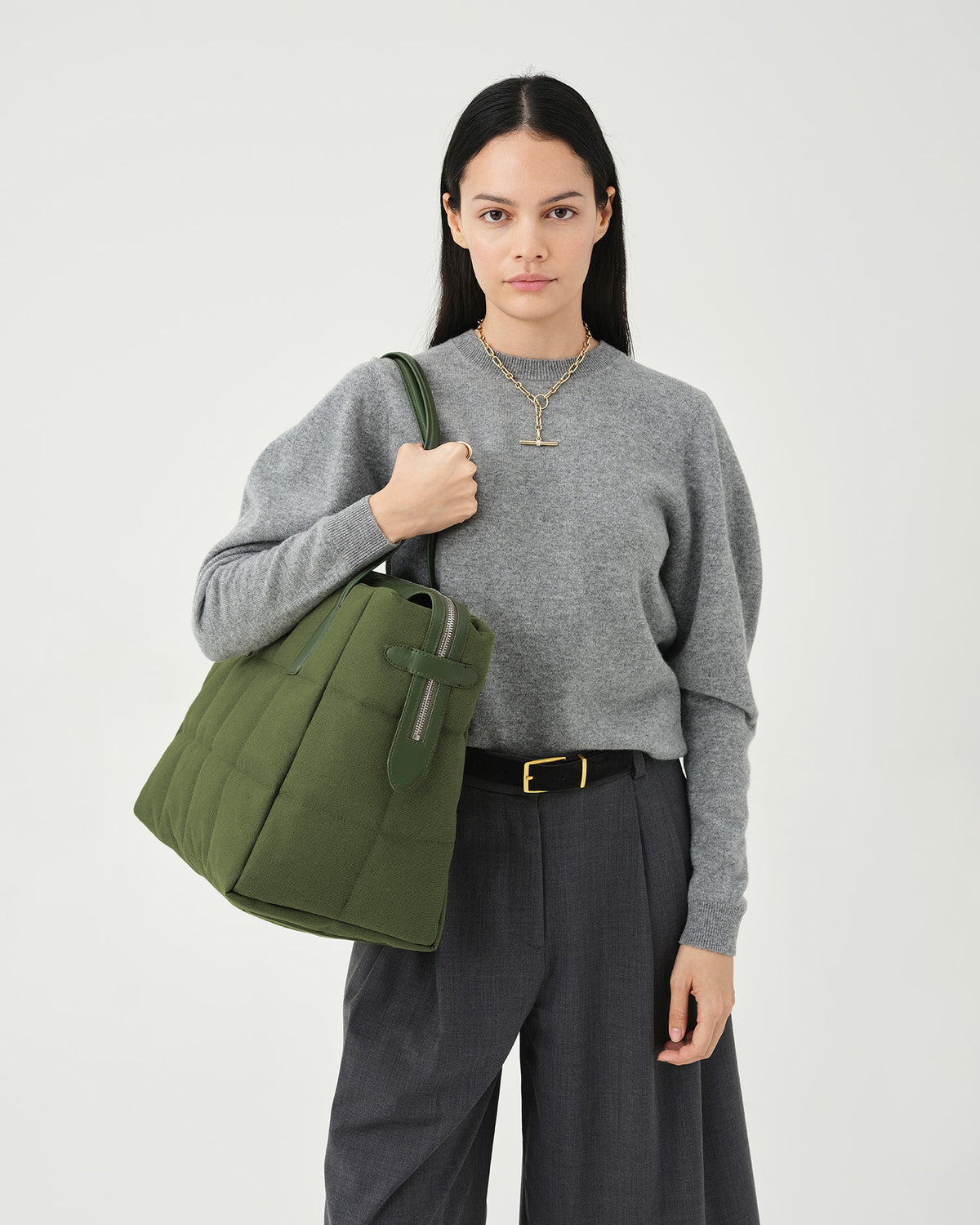 A photo of a woman modeling an olive Tela Tote bag with a matching shoulder strap, styled with a gray knit sweater, dark gray wide-leg trousers, against a plain white background.