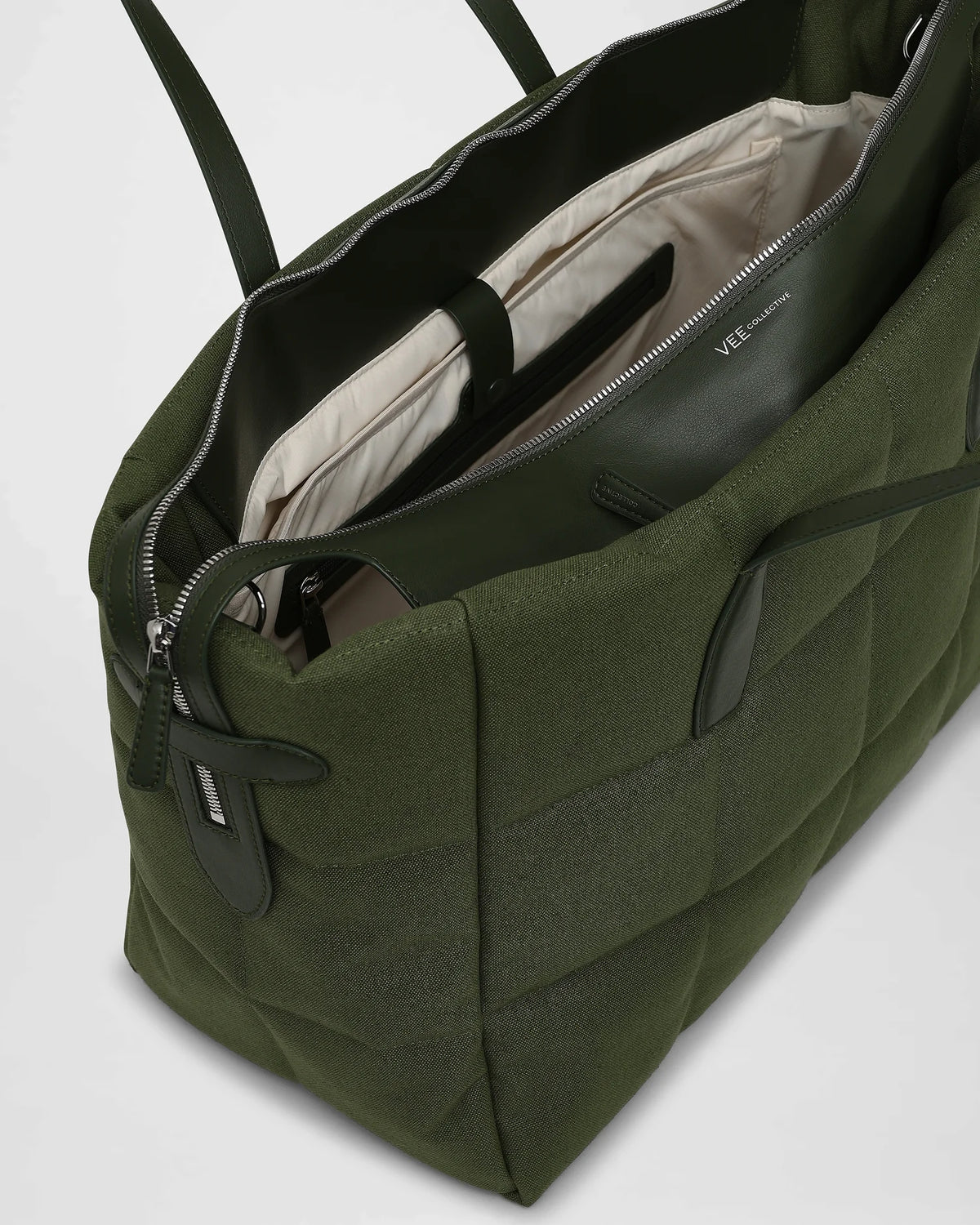 A picture featuring an open Tela Tote Bag in olive green with dark green leather trim and handles, revealing a cream-colored interior lining and a zippered pocket.