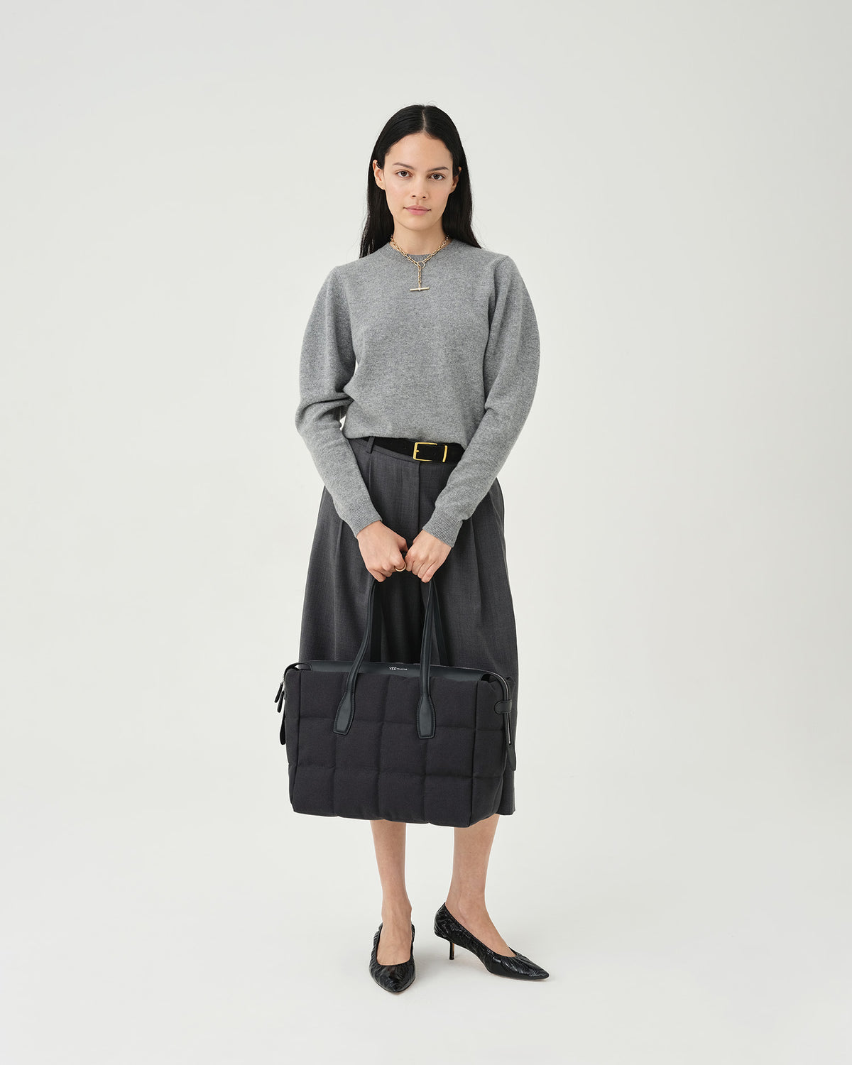 A photo of a model standing against a white studio background, wearing a gray long-sleeve knit top, dark charcoal pleated midi pants, and black pointed-toe heels, while carrying a Tela Tote with black handles.