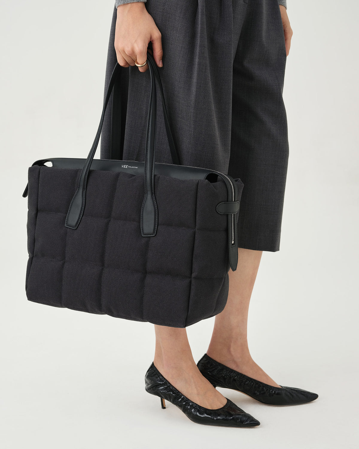 A photo of a model standing against a white studio background, wearing a gray long-sleeve knit top, dark charcoal pleated midi pants, and black pointed-toe heels, while carrying a Tela Tote with black handles.