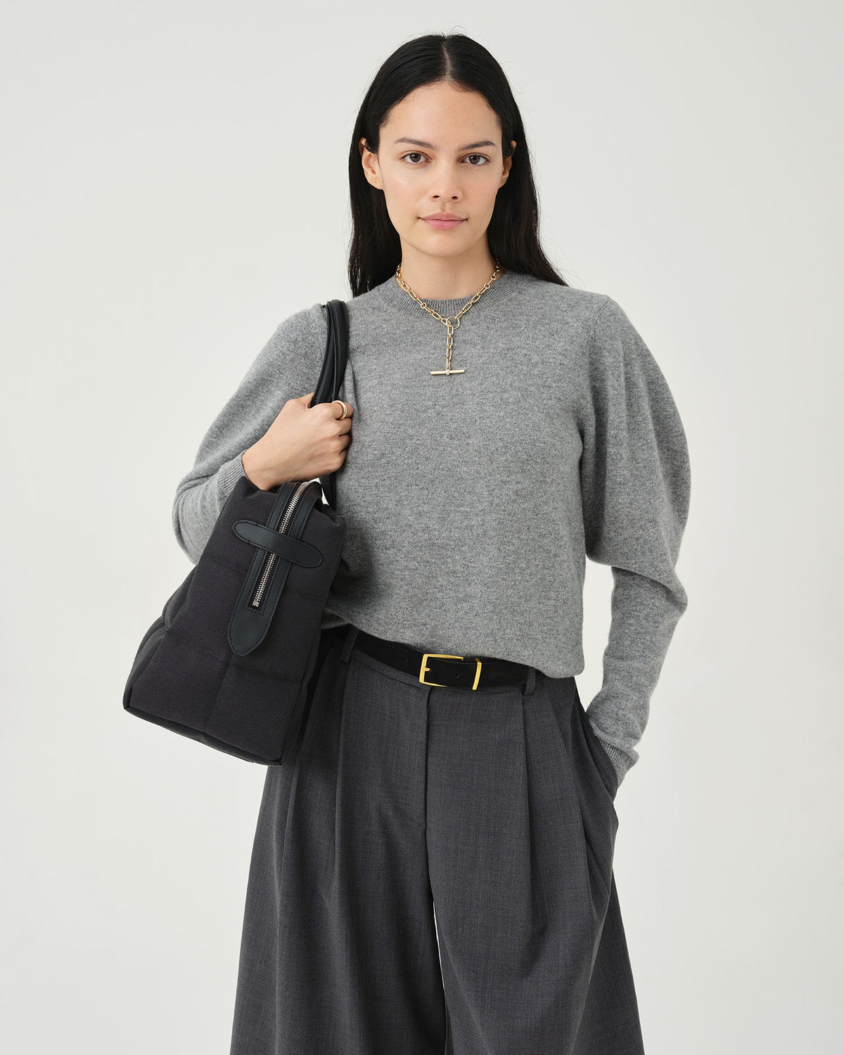 A photo of a model standing against a white studio background, wearing a gray long-sleeve knit top, dark charcoal pleated midi pants, and black pointed-toe heels, while carrying a Tela Tote with black handles.