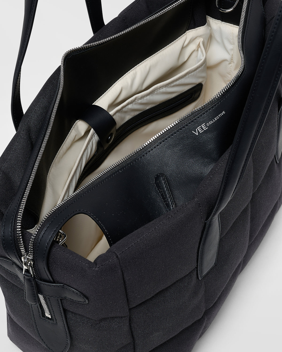 A close-up product photo of an open Tela Tote bag featuring black leather trim and interior lining, with a zippered compartment and interior pocket, while the “VEE” brand name is visible on the inner panel.