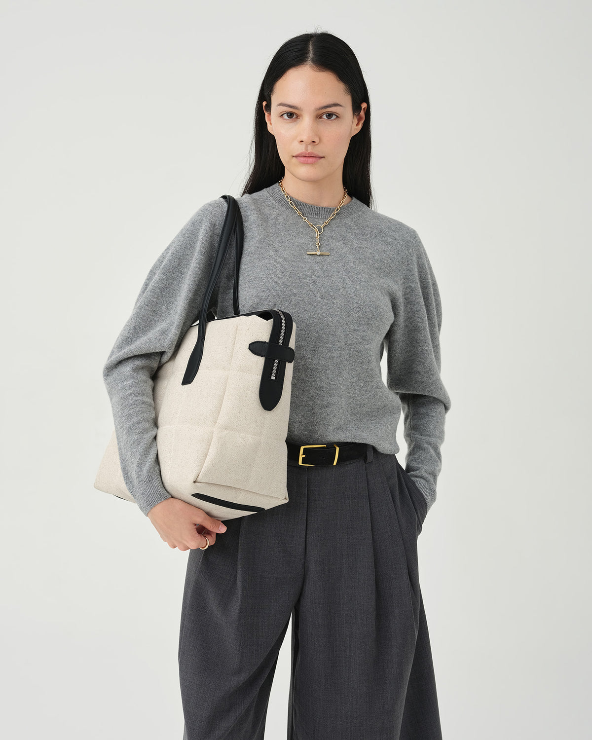 A photo of a model standing against a white studio background, wearing a gray long-sleeve knit top, dark charcoal pleated midi pants, and black pointed-toe heels, while carrying a Tela Tote with black handles.