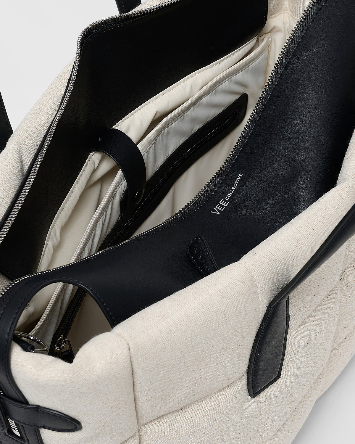 A close-up product photo of an open Tela Tote bag featuring black leather trim and interior lining, with a zippered compartment and interior pocket, while the “VEE” brand name is visible on the inner panel.