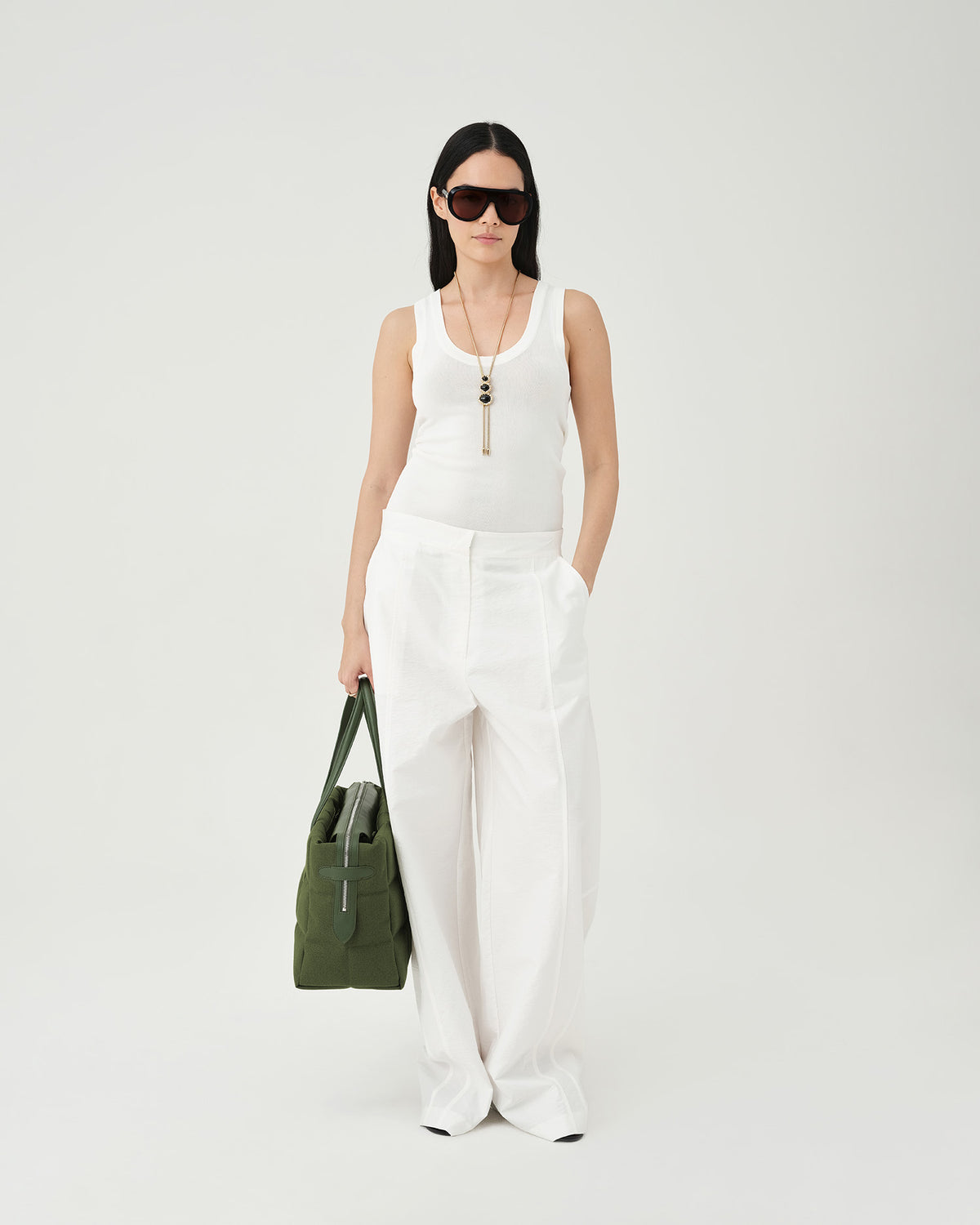 A photo of a woman wearing a white sleeveless top and white wide-leg pants, carrying olive Tela Tote shoulder bag against a plain light gray studio background.