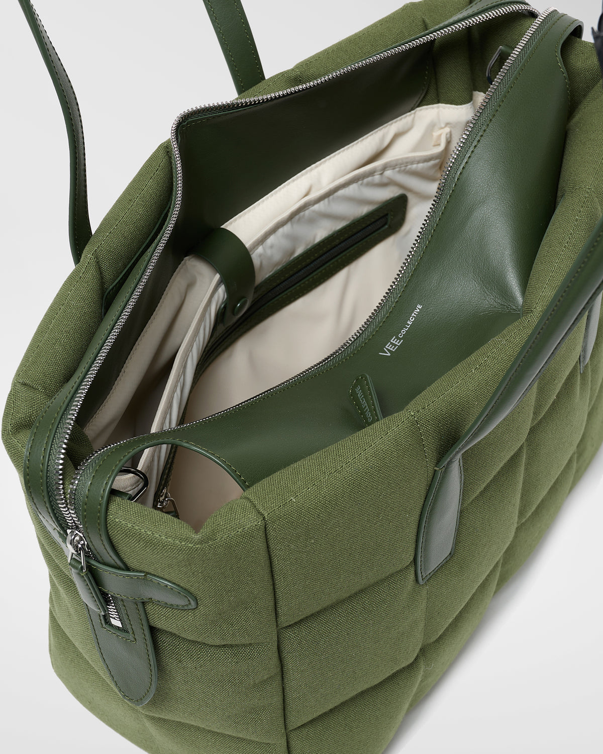 A close-up photo of an open Tela Tote in olive bag featuring black zippers and a light beige interior lining, set against a white background.