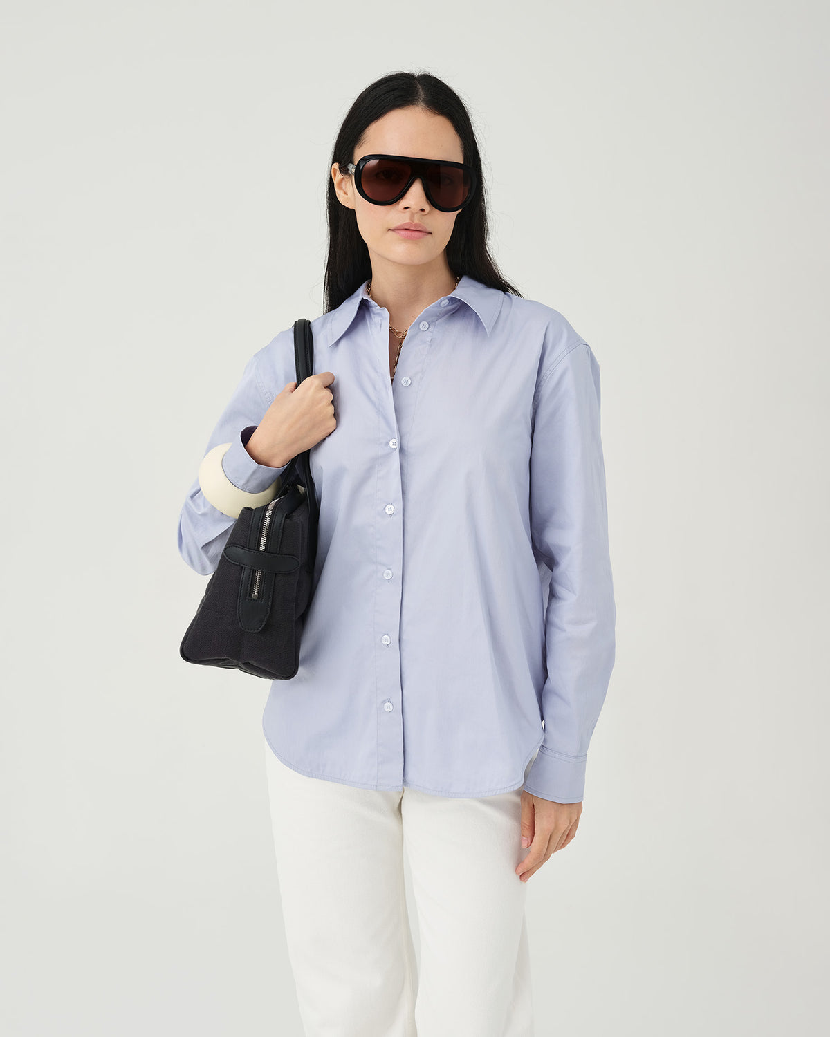 A studio fashion photo of a woman wearing a light blue button-up shirt and white trousers, carrying a Tela Tote bag set against a clean white background.