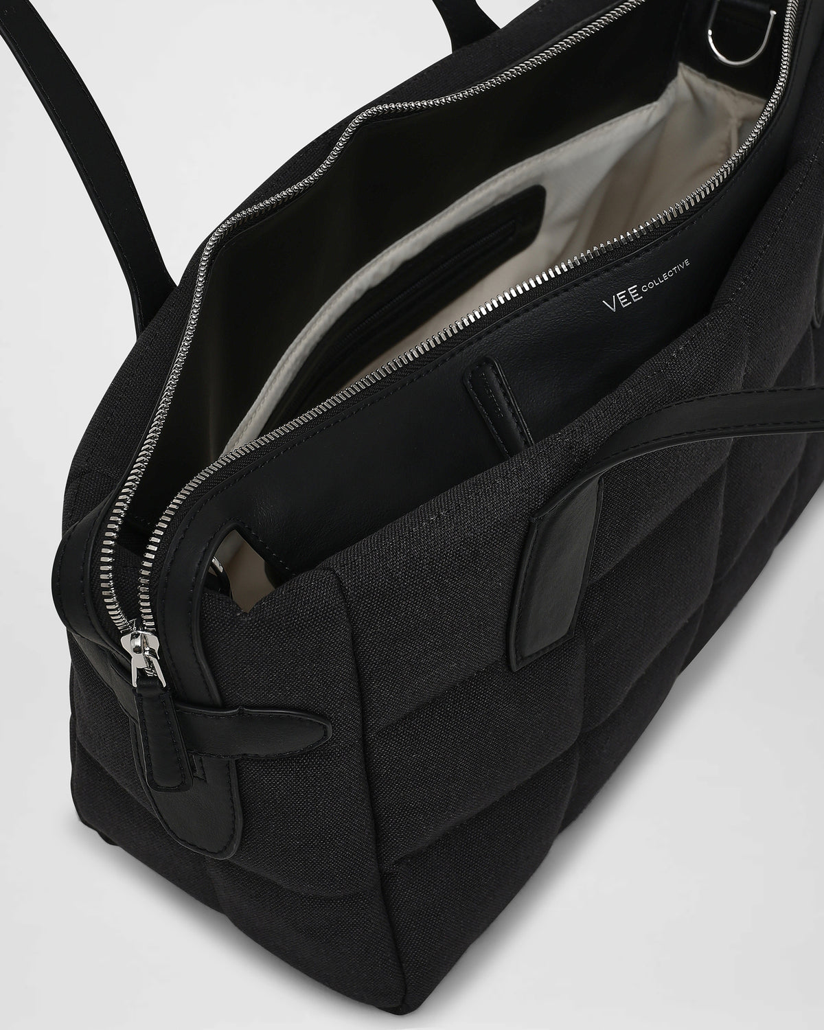 A close-up product photo of an open Tela Tote bag featuring black leather trim and interior lining, with a zippered compartment and interior pocket, while the “VEE” brand name is visible on the inner panel.