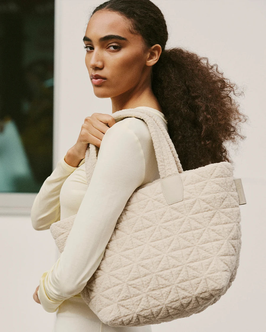 Side view of a woman’s upper body, wearing a Vee Tote bag Small cream sherpa by Vee Collective over her shoulder against a neutral background.
